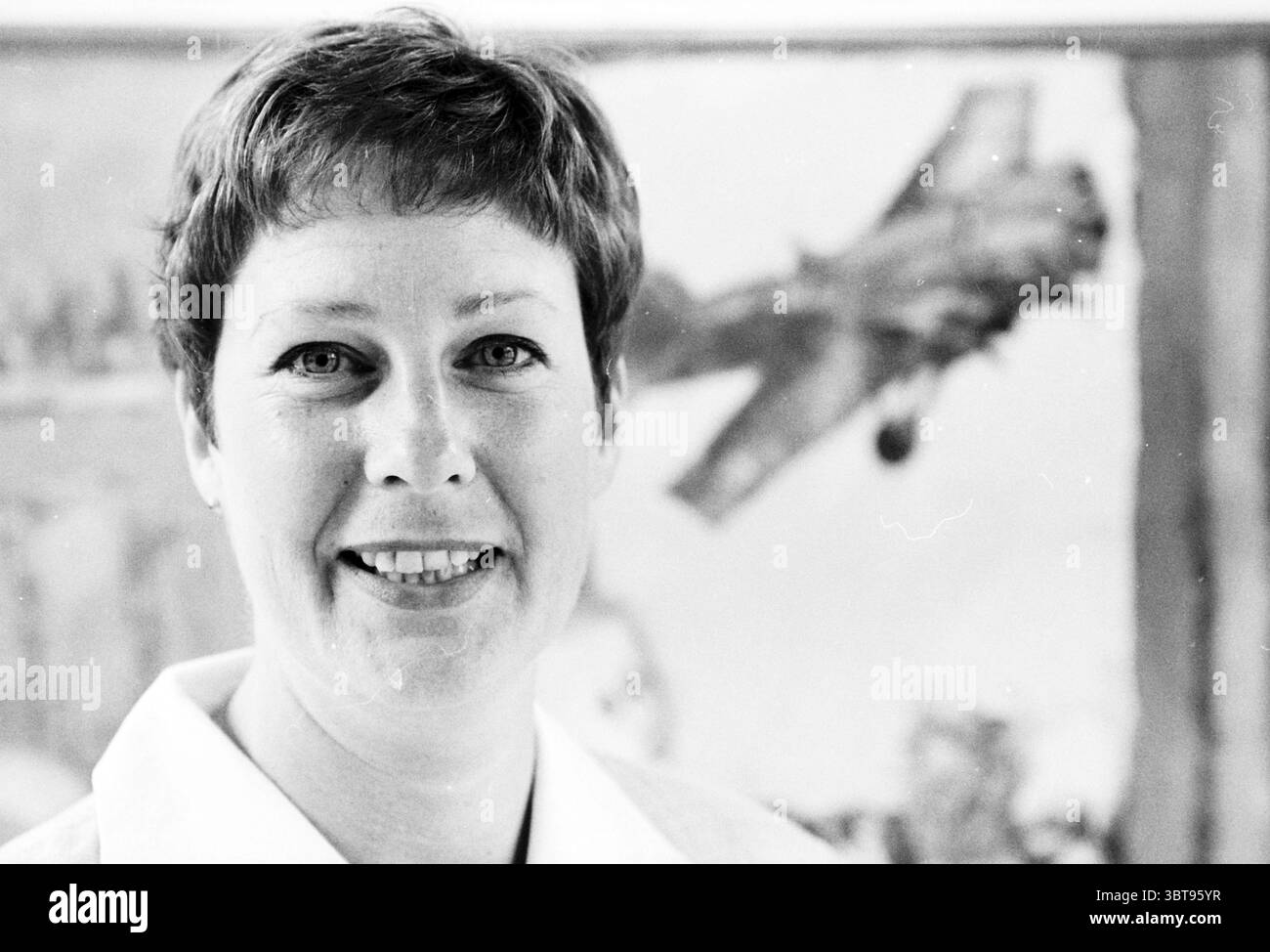 Janny v.d. Pluim Province of North Holland, Whizgle News, Dutch Desk, Niederlande, 1950 - 2000 am 09-07-1998. Die Abbildung zeigt diese Themen. In der Szene steht eine Frau mit kurzen, dunklen Haaren und einem fröhlichen Ausdruck im Vordergrund, ihr Gesicht wird von sanftem, natürlichem Licht beleuchtet. Sie trägt einen weißen Blazer, der einen Kontrast zu ihrem Hautton bildet und ihr offenes Auftreten unterstreicht. Ihre Augen sind hell und einnehmend, was Wärme und Freundlichkeit andeutet. Im Hintergrund zieht eine große, dynamische Darstellung eines Flugzeugs die Aufmerksamkeit auf sich. Das Flugzeug scheint in Aktion zu sein, vielleicht im Flug, und die Co Stockfoto