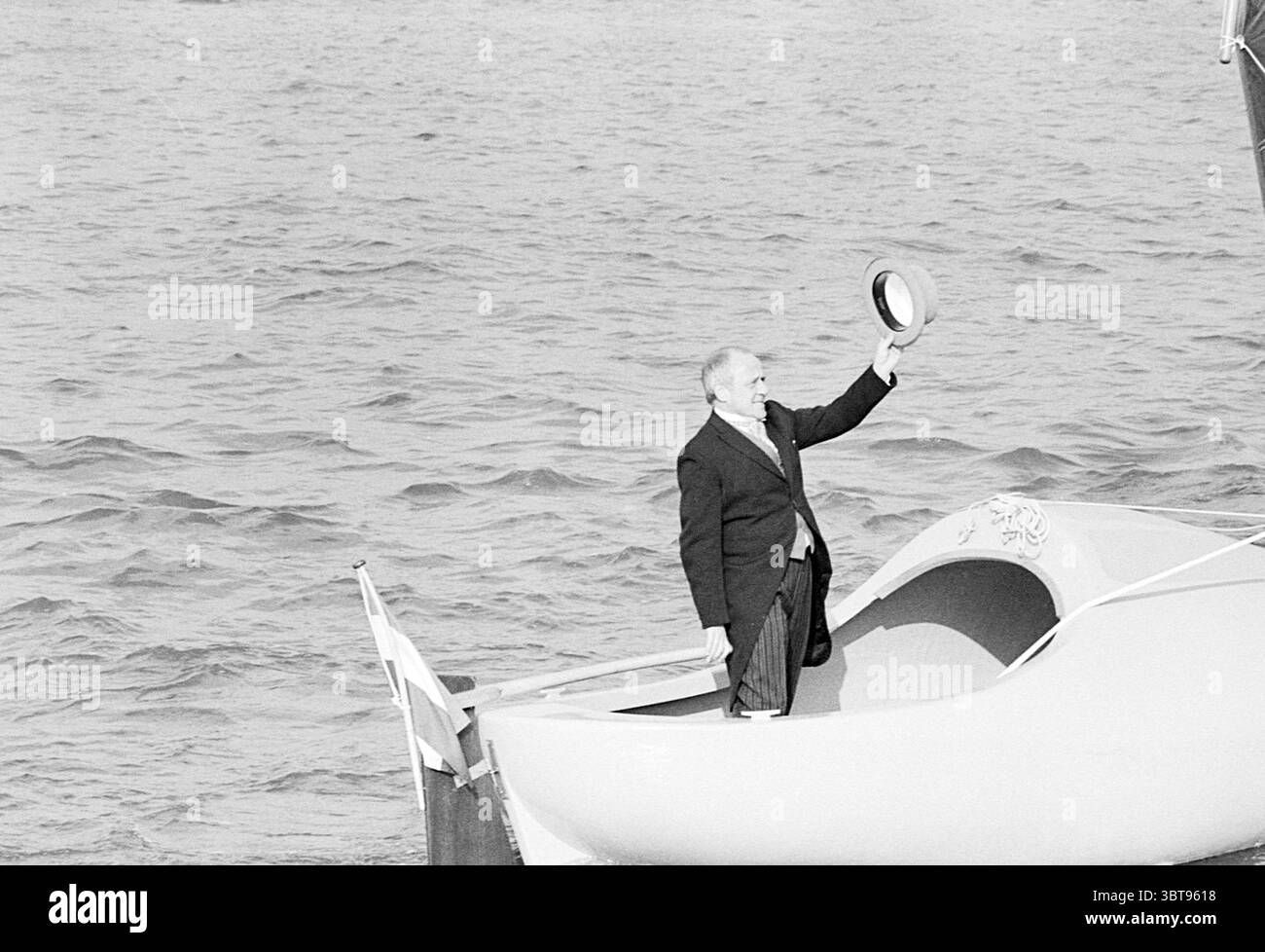 Einführung 'Klomp Jot' A'Dam von Johny Kraaykamp Schepen, Whizgle News, Dutch Desk, Niederlande, 1950 - 2000 am 07-03-1978. Diese Themen sind in der Abbildung dargestellt. In der Szene steht ein Mann in einem kleinen Boot, das auf dem Wasser driftet. Er trägt einen formellen Anzug, der einen Hauch von Eleganz suggeriert, und er hält mit der rechten Hand einen Strohhut hoch, als würde er andere begrüßen oder sich von anderen verabschieden. Das Boot selbst ist einfach, aber gut gestaltet, mit einem sanft geschwungenen Rumpf und einer eingebetteten Seite, die ein Gefühl von Auftrieb verleiht. Die Oberfläche des Wassers ist hügelig, reflektiert Licht in glitzernden Mustern, und das ov Stockfoto