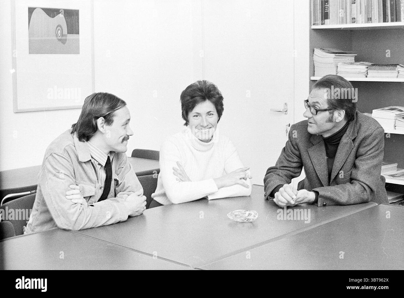 Cultural Council NH Persons, Whizgle News, Dutch Desk, Niederlande, 1950 - 2000 am 06-10-1971. Das Bild enthält diese Themen. In einem gut beleuchteten, minimalistischen Raum mit einem hellen, neutralen Farbschema sitzen drei Personen um einen eleganten, dunklen Tisch. Die Umgebung scheint eine Atmosphäre der Zusammenarbeit oder des Gesprächs zu vermitteln, die ein professionelles, aber entspanntes Umfeld widerspiegelt. Links lehnt sich ein Mann mit mittellangen Haaren, der lässig in einer leichten Jeansjacke über einem schlichten Hemd gekleidet ist, leicht nach vorne, lächelt und mit den anderen anspricht. Er hat ein warmes, offenes Auftreten, das ihn andeutet Stockfoto