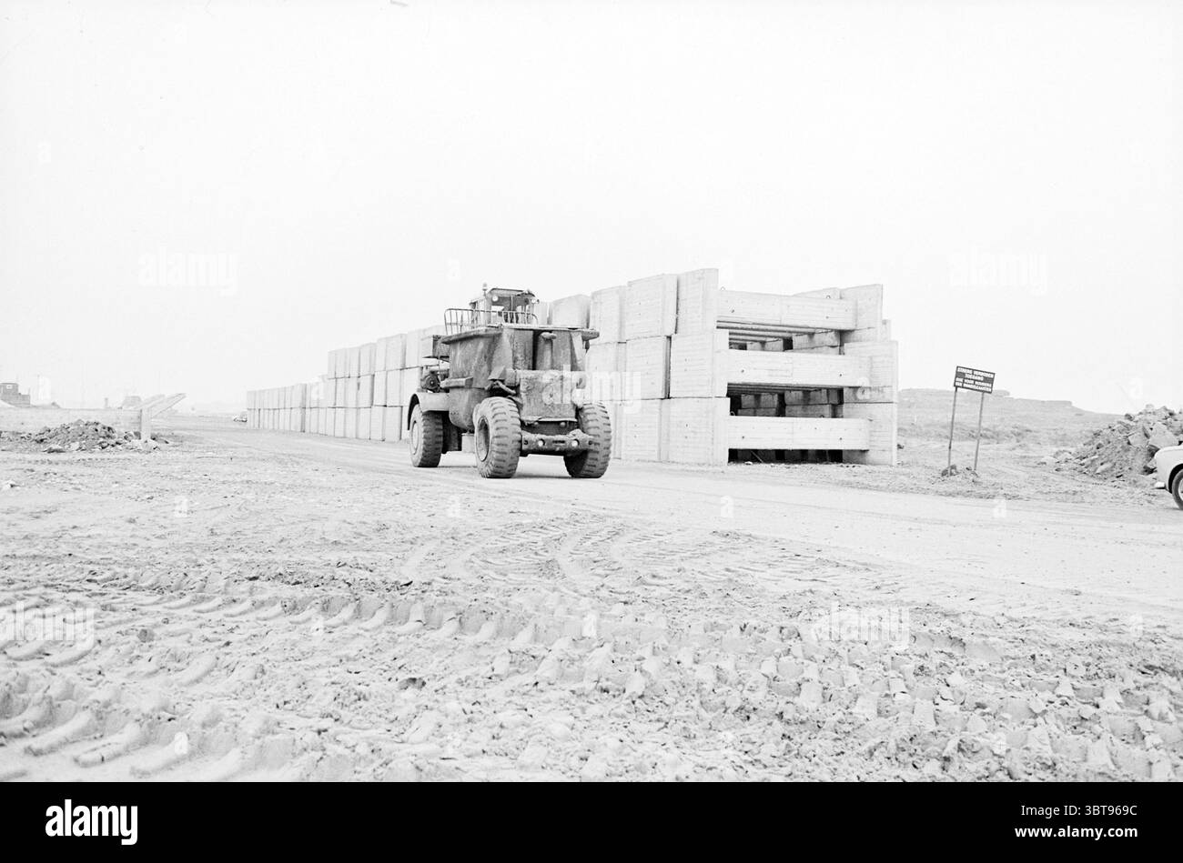 Bulldozer at Concrete Elements., Whizgle News, Dutch Desk, Niederlande, 1950 - 2000. Dies sind die Elemente im Bild. Die Szene fängt eine riesige, offene Baustelle unter einem bewölkten Himmel ein und schafft eine gedämpfte, bewölkte Atmosphäre. Der Boden ist ein Flickenteppich aus lockerer, gestörter Erde, die mit tiefen Reifenspuren markiert ist, die auf kürzliche Aktivitäten hinweisen. Im Vordergrund dominiert ein großes Baufahrzeug die Szene, dessen robustes Äußere Verschleißspuren mit einer Staubschicht zeigt, was darauf hindeutet, dass es unermüdlich gearbeitet hat. Die massiven Räder vermitteln Kraft und Bewegung, während das Fahrzeug in seiner Position steht Stockfoto