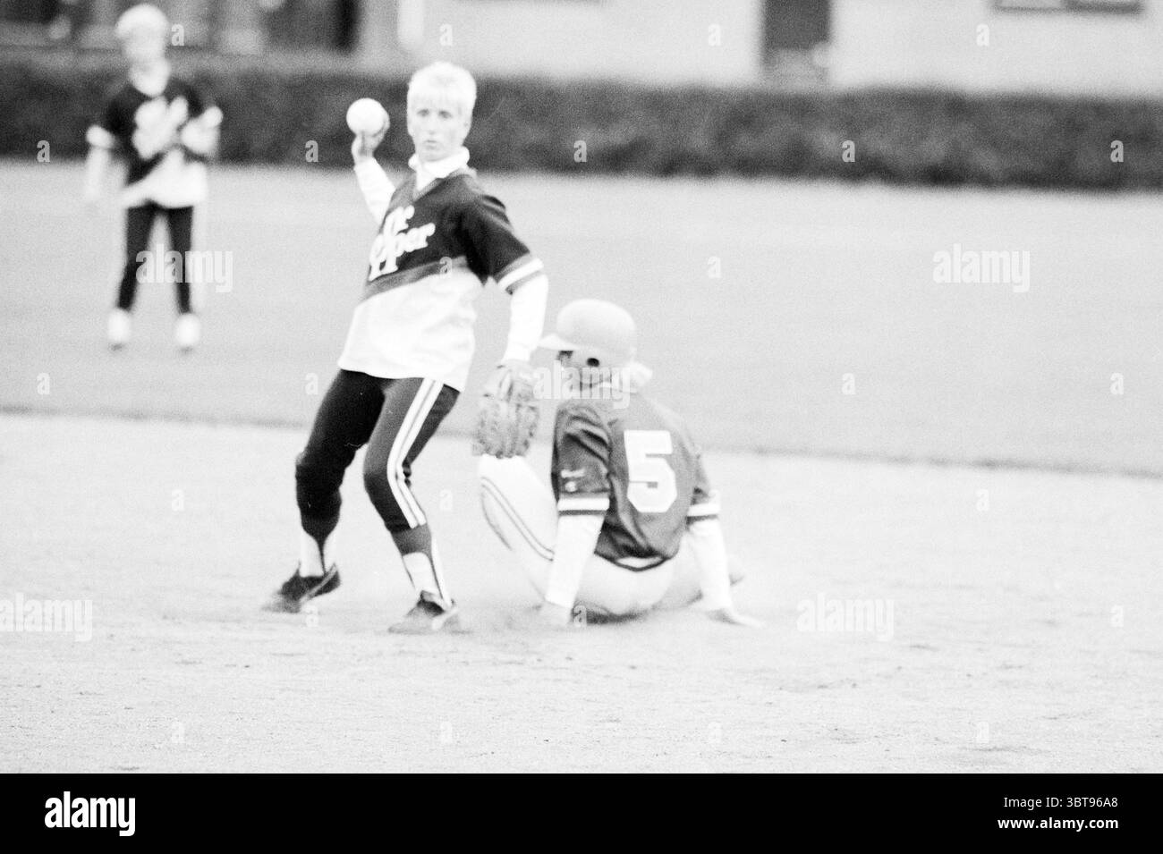 HHC - Heiloo Softball, Whizgle News, Dutch Desk, Niederlande, 1950 - 2000 am 12-08-1987. Dies sind die Elemente im Bild. Die Szene fängt einen Moment auf einem Baseballfeld während eines Spiels ein. Im Vordergrund steht ein Spieler in einem hellen Trikot mitten im Wurf und zeigt eine athletische Haltung. Ihr Ausdruck ist bestimmt, und sie haben kurze, helle Haare. Dieser Spieler trägt schwarze Leggings, die mit weißen Streifen verziert sind und für ein dynamisches Flair sorgen. Auf dem Boden sitzt ein weiterer Spieler, der in einem hellroten Trikot gekleidet ist, wobei die Nummer 5 deutlich sichtbar dargestellt wird. Sie sind in einer defensiven Position, Stockfoto