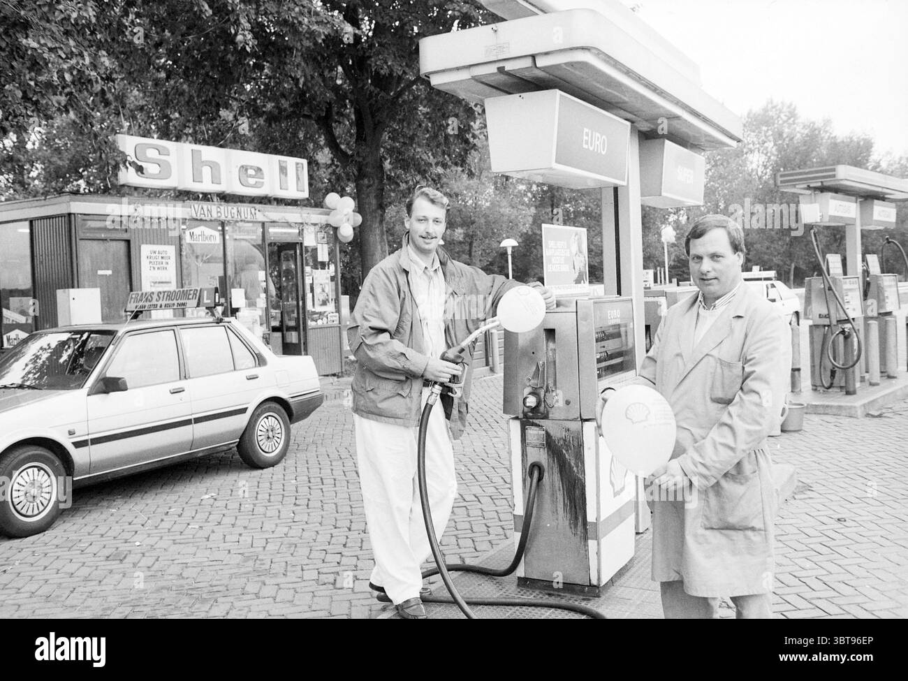 Zwei Männer in der Shell Van Bugnum Pumpstation in Santpoort Santpoort, Whizgle News, Dutch Desk, Niederlande, 1950 - 2000 am 02-10-1989. Dies sind die Themen im Bild. Die Szene zeigt zwei Männer, die an einer Tankstelle stehen und weiße Ballons halten. Sie befinden sich neben einer Kraftstoffpumpe, die über ein großes, übersichtliches Display mit dem Preis in Euro verfügt. Die Männer tragen helle Kleidung, einer trägt einen hellen Blazer und eine Hose, der andere trägt einen beigen Trenchcoat. Ihre Ausdrucksformen wirken fröhlich und deuten auf eine feierliche Atmosphäre hin. Die Tankstelle selbst hat ein Rev Stockfoto