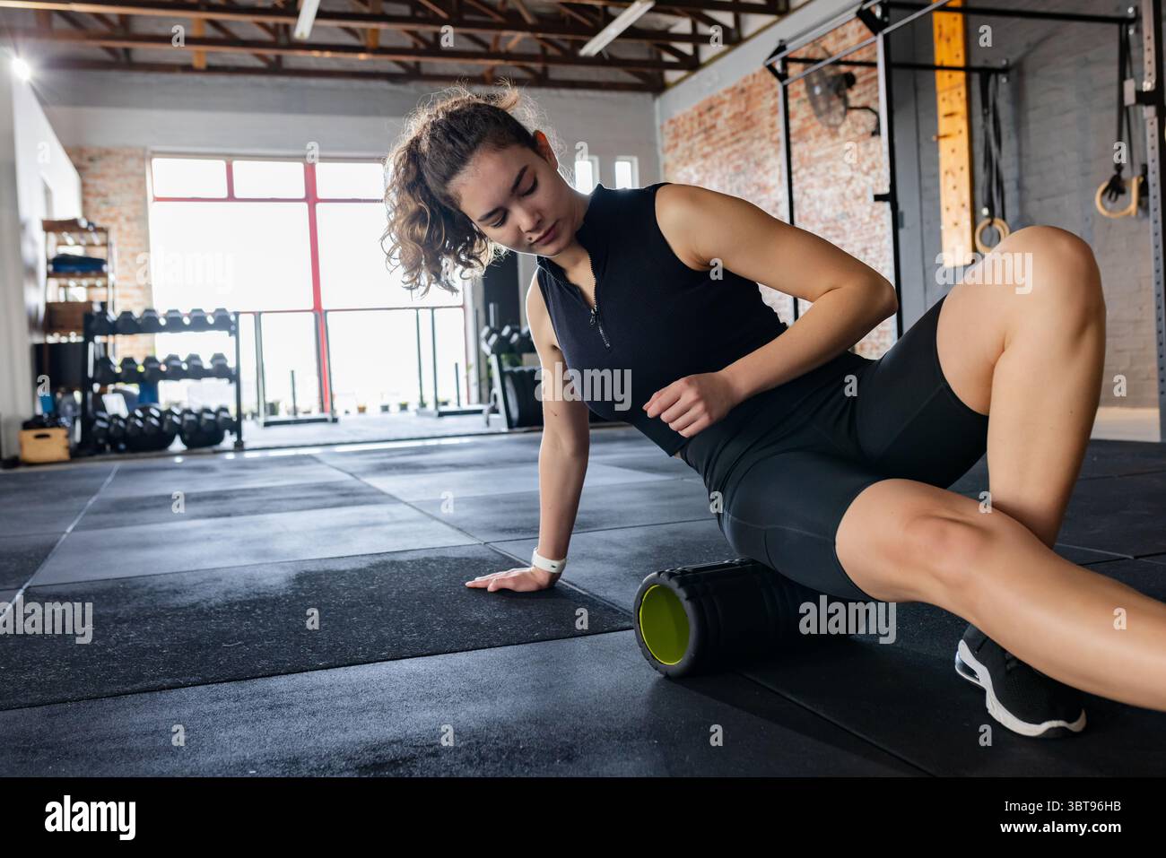 Erwachsene weibliche Fitness-Enthusiasten sitzen auf einer Turnmatte mit Schaumstoffrolle am Oberschenkel in der Nähe des Hantelhalters Stockfoto