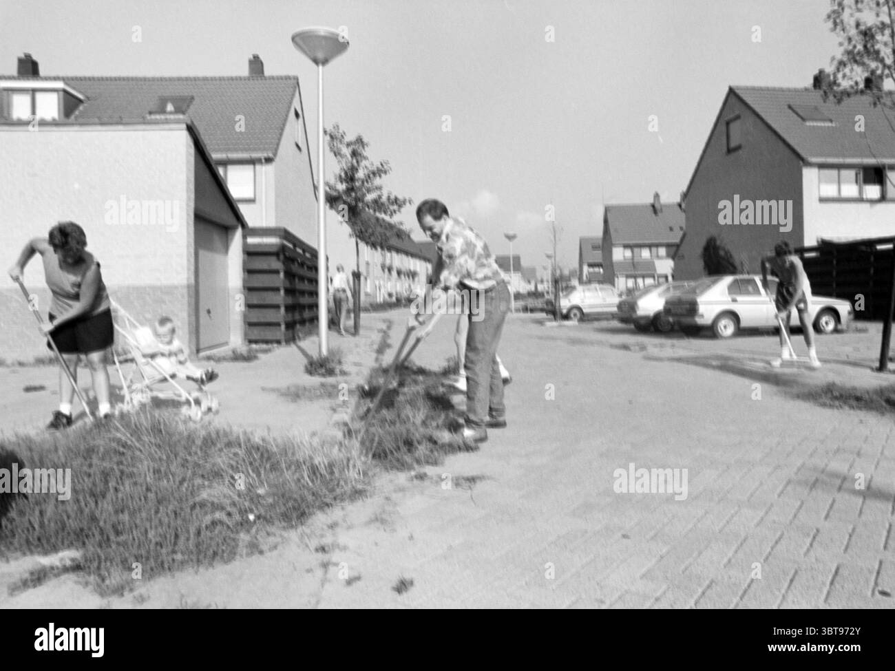 Entfernen Sie Unkräuter Velserbroek, Whizgle News, Dutch Desk, Niederlande, 1950 - 2000 am 26-08-1990. Diese Themen sind in der Abbildung dargestellt. In dieser Szene wird eine Wohnstraße dargestellt, die von engen Häusern gesäumt ist, deren Fassaden hauptsächlich in gedämpften Grau- und Weißtönen gehalten sind. Die Architektur verfügt über Giebeldächer, die sich durch ein einfaches und funktionales Design auszeichnen. Eine Gruppe von drei Personen beschäftigt sich mit Gartenarbeiten, die sich alle auf das Beseitigen von bewachsenem Gras und Unkraut konzentrieren. Eine Person, die in legere Kleidung gekleidet ist, beugt sich mit einem Werkzeug nach unten und schneidet aktiv die Vegetation ab. Nach links, eine weitere Figur Stockfoto