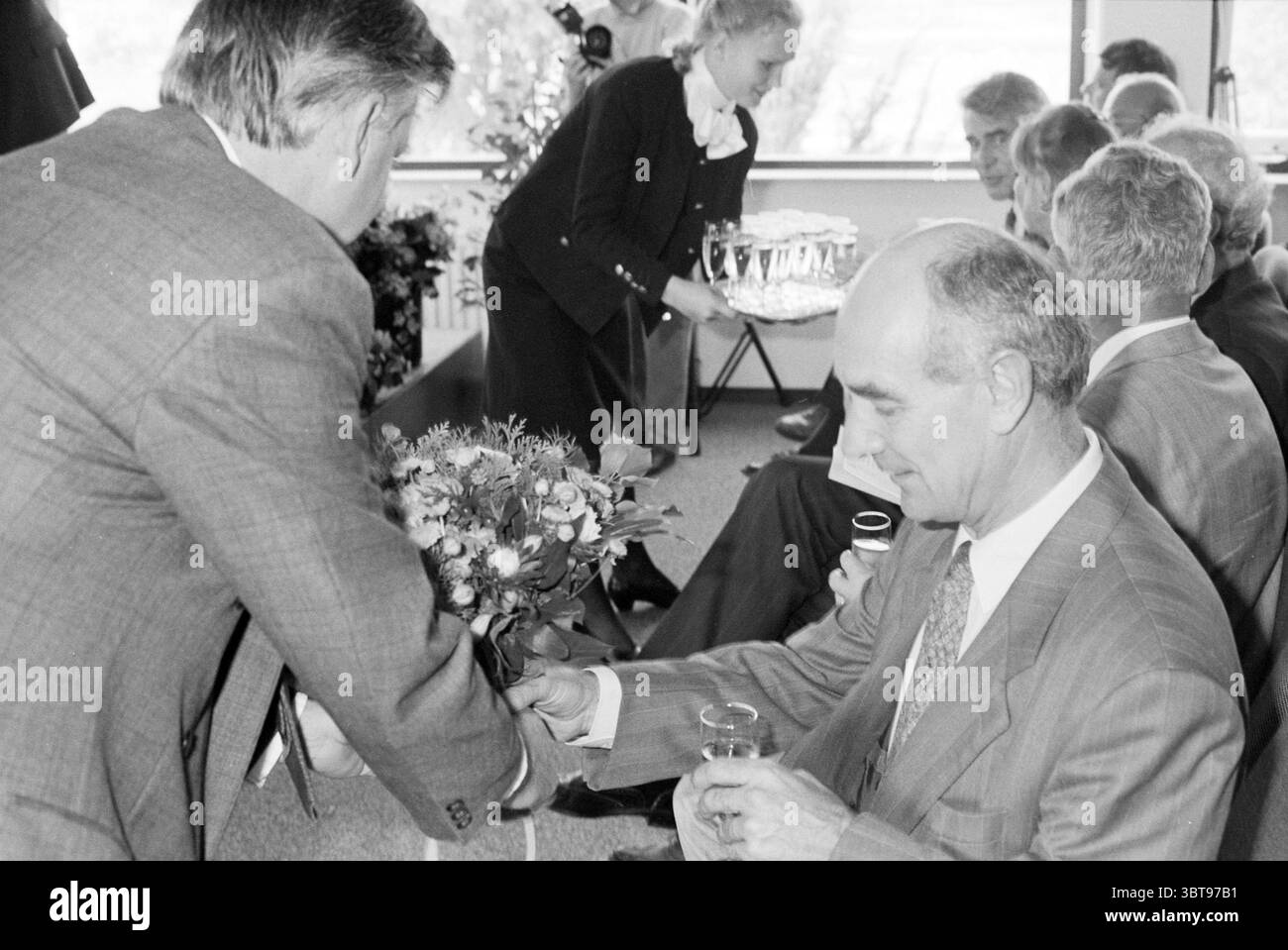 Minister Klaas de Vries eröffnet BGD, Whizgle News, Dutch Desk, Niederlande, 1950 - 2000 am 07.09.1993. Dies sind die Themen im Bild. In einem hellen, luftigen Innenbereich findet ein geselliges Treffen statt. Die Atmosphäre vermittelt ein Gefühl von Feier oder Formalität. Auf der einen Seite streckt ein Mann in einem hellen Anzug einen Blumenstrauß zu einer anderen Person, die lächelt und sitzt. Dieser sitzende Mann, in einem dunkleren Anzug gekleidet, scheint engagiert und dankbar für die Geste zu sein. Die Blumen sind lebendig und kontrastieren mit den gedämpften Tönen der Anzüge, was die festliche Atmosphäre des unterstreicht Stockfoto