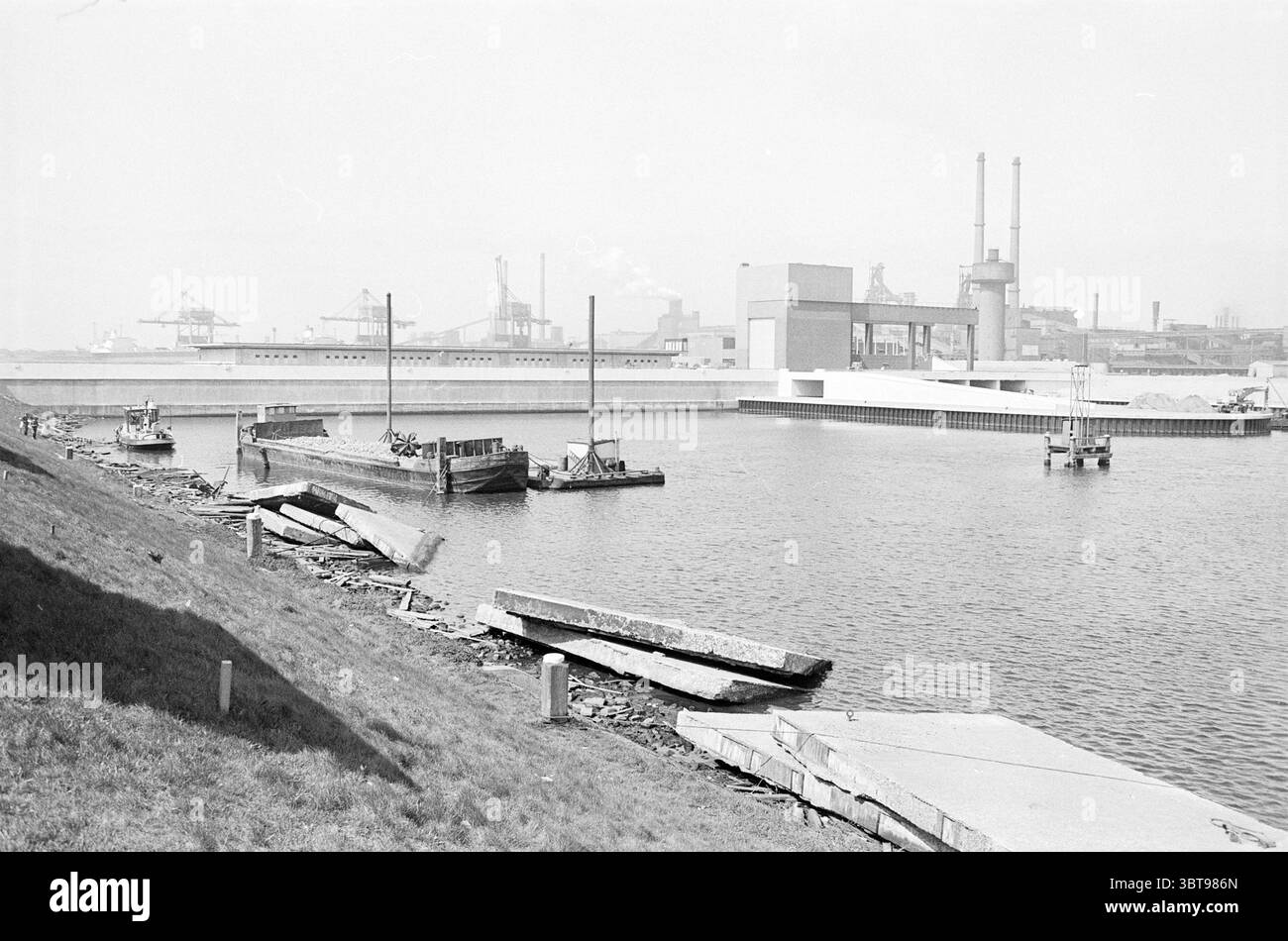 Spuigemaal in IJmuiden Waterworks, Whizgle News, Dutch Desk, Niederlande, 1950 - 2000 am 18-04-1974. Dies sind die Themen im Bild. Die Szene fängt eine ruhige und dennoch industrielle Uferpromenade ein, umgeben von einer sanften, gedämpften Grau- und Weißpalette. Das Wasser ist ruhig und reflektiert den bewölkten Himmel, während es sanft an den Rändern entlang weht, wo es auf das Land trifft. Links sind mehrere Boote vertäut, deren Formen gegen das Wasser geschildert sind. Sie scheinen verwittert zu sein, was auf ihren langen Dienst hindeutet und sind mit dem industriellen Charakter des Ortes verbunden. Stockfoto