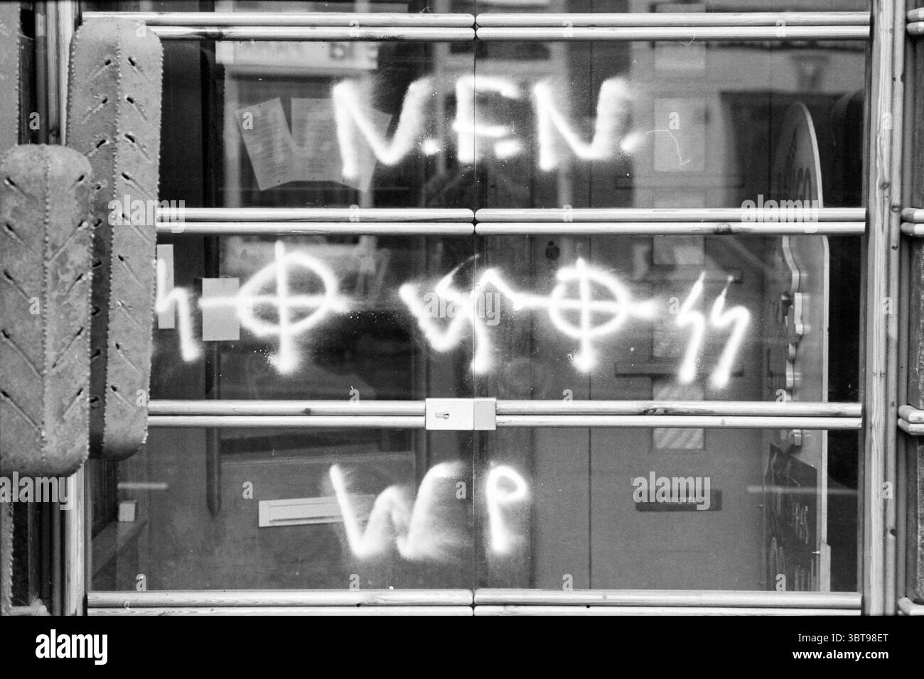Rassisten-Slogans Rest, Whizgle News, Dutch Desk, Niederlande, 1950 - 2000 am 16-08-1994. Diese Themen werden in der Abbildung angezeigt. Die Szene zeigt ein Schaufensterfenster, das stark von Graffiti geprägt ist und sich stark von der Glasoberfläche abhebt. Das Graffiti besteht aus mehreren Symbolen und Buchstaben, die in weißer Farbe über das Fenster gestreut sind und einen scharfen Kontrast zum klaren Glas dahinter bilden. Der Fensterrahmen ist metallisch, mit einem Gitter aus horizontalen und vertikalen Balken, die den Raum in Abschnitte unterteilen. Die Struktur des Rahmens ist leicht abgenutzt, was auf Alter oder Witterungseinflüsse hindeutet. Hinter dem Fenster, schwach sil Stockfoto