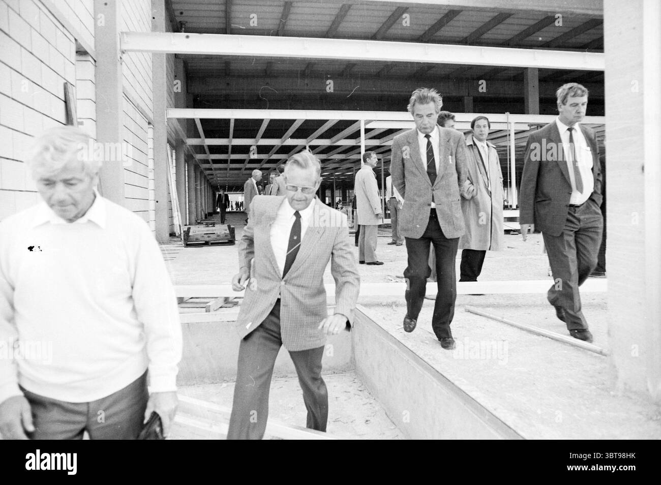 Höchster Punkt C & A Lisse höchster Punkt Lisse Kanaalstraat, Whizgle News, Dutch Desk, Niederlande, 1950 - 2000 am 07-08-1986. Diese Themen werden in der Abbildung angezeigt. In der Szene läuft eine Gruppe gut gekleideter Männer durch eine unvollendete Baustelle. Die Anlage verfügt über eine große offene Fläche mit Stahlträgern und Holzkonstruktionen, die auf eine laufende Entwicklung hindeuten. Die Männer tragen formelle Kleidung; einige tragen Anzüge und Krawatten, während eine Person in einem hellen Pullover auffällt. Die Texturen ihrer Kleidung stehen im Kontrast zur rauen, industriellen Umgebung. Stockfoto