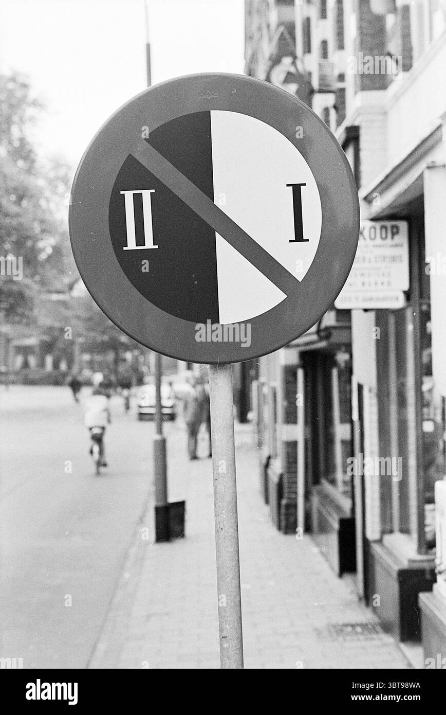 Verkehrsschilder Beverwijk Verkehrssicherheit, Whizgle News, Dutch Desk, Niederlande, 1950 - 2000 am 29-08-1967. Das Bild enthält diese Themen. Die Szene fängt eine urbane Straße ein, wahrscheinlich in einer belebten Stadtumgebung. Im Vordergrund befindet sich ein rundes Verkehrsschild mit deutlichen schwarzen und weißen Segmenten, das eine auffällige diagonale Linie zeigt, die auf ein Verbot oder eine Einschränkung hinweist. Die visuelle Spannung, die durch die kontrastfarbenen Farben erzeugt wird, lenkt die Aufmerksamkeit und unterstreicht die Bedeutung der Botschaft, die durch das Zeichen vermittelt wird. Im Hintergrund befinden sich Gebäude mit einer Mischung aus architektonischen Stilen Stockfoto