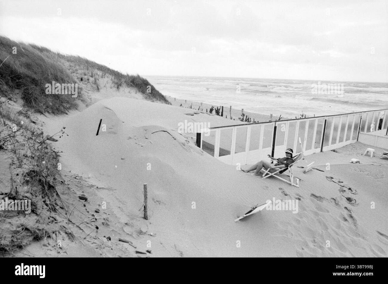 Stiffed Sand Zandvoort Sand Zandvoort, Whizgle News, Dutch Desk, Niederlande, 1950 - 2000 am 05-08-1985. Die Abbildung zeigt diese Themen. Die Szene zeigt eine Küstenlandschaft, die von sandigen Hügeln geprägt ist, was auf windgepeitschte Dünen hinweist. Die Sandkörner in verschiedenen hellen Beige- bis Grau-Tönen erzeugen eine weiche, wellige Textur, die sich nahtlos mit dem Horizont verschmelzen lässt. Auf der einen Seite steht ein hölzernes Geländer, teilweise verdeckt durch den driftenden Sand. Die verwitterte, weiße Farbe deutet auf eine lange Exposition gegenüber den Elementen hin und kontrastiert mit den dunkleren Tönen der san Stockfoto