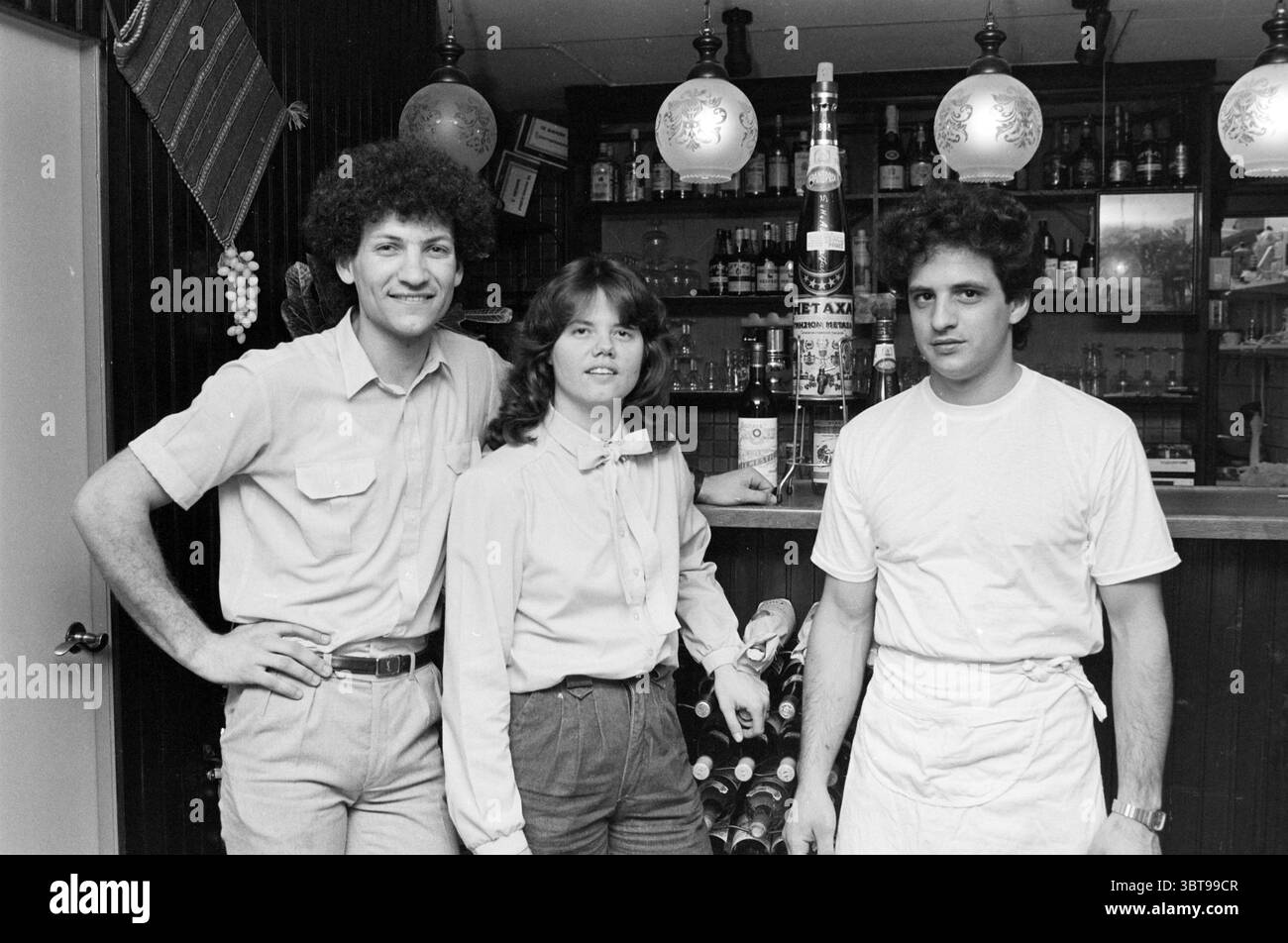 Neues griechisches Restaurant Zorba The Greek Restaurant, Whizgle News, Dutch Desk, Niederlande, 1950 - 2000 am 15-05-1985. Dies sind die Elemente im Bild. In einer gemütlichen Bar stehen drei Personen zusammen und verströmen eine freundliche Atmosphäre. Die Kulisse ist mit dunkler Holztäfelung geschmückt, die im Kontrast zum warmen Leuchten hängender Pendelleuchten steht. Diese Lichter sorgen für eine sanfte Beleuchtung, die die Regale der Bar mit verschiedenen Flaschen hervorheben und ein Gefühl von Tiefe und Reichtum im Raum schaffen. Die erste Person links, die ein helles kurzärmliges Hemd trägt, hat Volumen Stockfoto