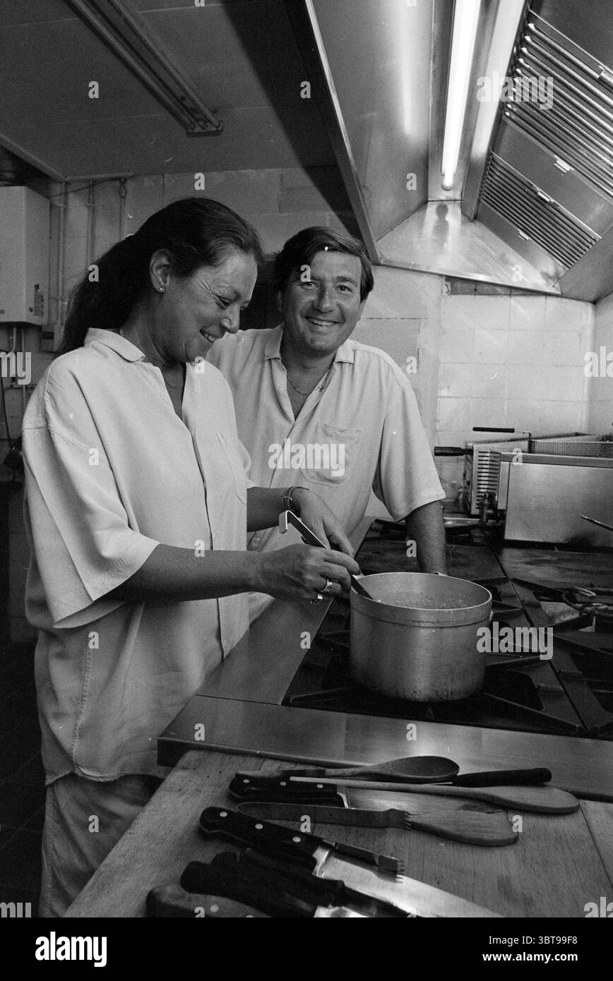 Chefrestaurant Pimpernel Hoofddorp Niederlande, Whizgle News, Dutch Desk, Niederlande, 1950 - 2000 am 03-05-1990. Das Bild enthält diese Themen. Die Szene zeigt eine geschäftige Küchenumgebung, die von Aktivität und Wärme lebhaft ist. Zwei Personen stehen an einem Edelstahlofen und sind tief in den Kochprozess eingebunden. Die Frau, mit langen Haaren nach hinten gezogen, lächelt, während sie einen Topf auf dem Brenner rührt, ihre Haltung entspannt und konzentriert. Sie trägt ein helles, locker geschnittenes Hemd, das für die Wärme der Küche geeignet ist. Neben ihr lehnt sich ein Mann an die Arbeitsplatte, ebenfalls lässig gekleidet Stockfoto