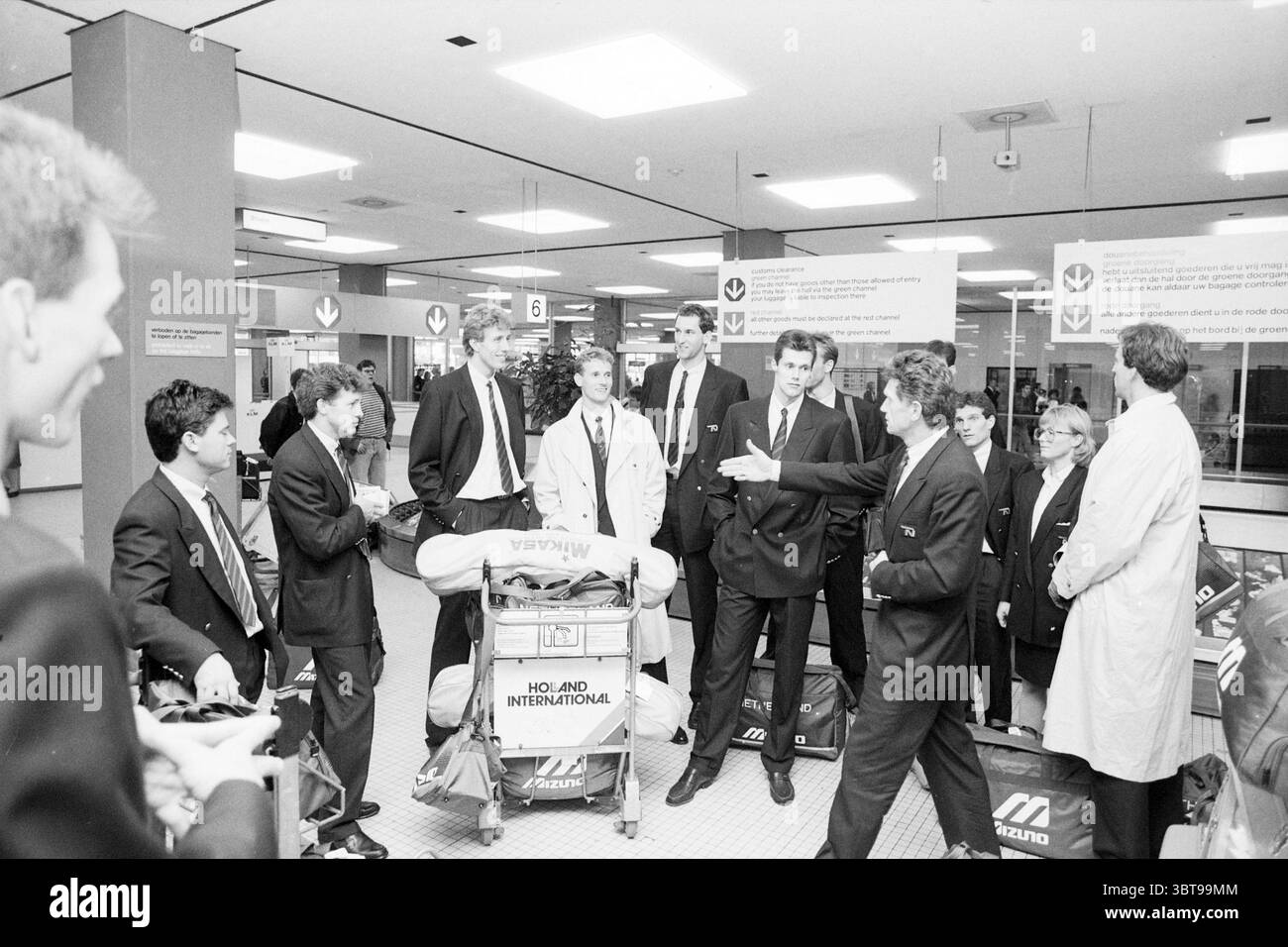 Ankunft der Volleyballmannschaft bei Schiphol Volleyball Schiphol, Whizgle News, Dutch Desk, Niederlande, 1950 - 2000 am 01-10-1989. Dies sind die Elemente im Bild. In einem geschäftigen Flughafenterminal versammeln sich eine Gruppe von Einzelpersonen um eine Reihe von Gepäckwagen, von denen jeder mehrere Taschen mit verschiedenen Logos hält. Die Szene ist monochromatisch, wobei Grautöne dominieren, die eine ernste und formale Atmosphäre widerspiegeln. Die Menschen sind in formeller Kleidung gekleidet; dunkle Anzüge und Krawatten sind bei den Männern vorherrschend, während mindestens ein Individuum in einem weißen Mantel auffällt, was auf ein mögliches hindeutet Stockfoto