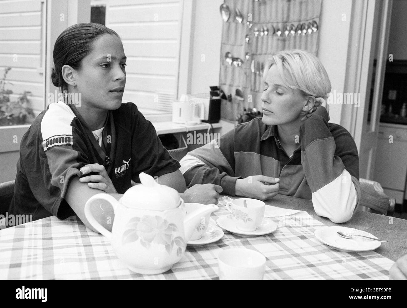 Zwei Wasserpolo-Spieler Haarlem the Netherlands, Whizgle News, Dutch Desk, Niederlande, 1950 - 2000 am 26.06.1996. Dies sind die Elemente im Bild. In einem gemütlichen Innenbereich sitzen zwei junge Frauen an einem Tisch. Das eine hat lange, dunkle Haare, die in einem eleganten Stil zurückgezogen werden und ein lässiges schwarz-weißes Hemd tragen, während das andere kurze, hellblonde Haare hat und ein farbenfrohes, gestreiftes Oberteil in Blau-, Rot- und Weißtönen trägt. Die Atmosphäre ist intim und reflektierend und vermittelt ein Gefühl tiefer Kontemplation oder Nachdenklichkeit. Der Tisch ist mit einer karierten Tischdecke in hellen Tönen und geschmückt Stockfoto