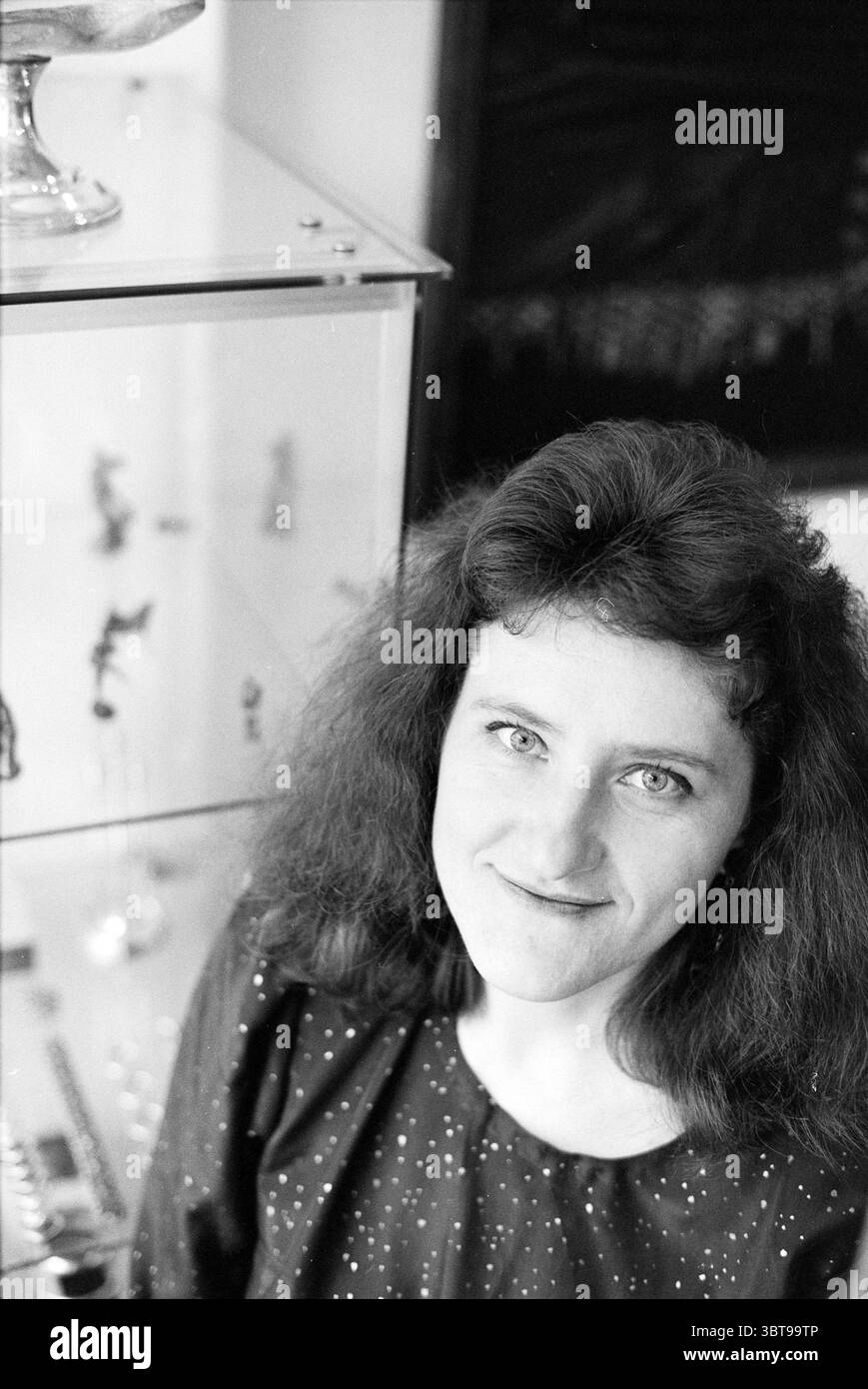 Porträt einer Frau, Whizgle News, Dutch Desk, Niederlande, 1950 - 2000 am 06-1991. Dies sind die Elemente im Bild. In dieser visuellen Szene posiert eine Frau mit lockigen dunklen Haaren direkt zum Betrachter. Ihr Ausdruck ist ruhig und leicht rätselhaft, mit einem Hauch eines Lächelns auf ihren Lippen. Sie trägt ein dunkles Kleid mit einem dezenten Muster aus kleinen, hellen Flecken, die ihrem Aussehen einen Hauch von Zartheit verleihen. Im Hintergrund befindet sich ein Vitrinenschrank mit verschiedenen Gegenständen, wahrscheinlich dekorativen Gegenständen oder Sammlerstücken, die einen persönlichen oder künstlerischen Raum widerspiegeln. Das cabi Stockfoto