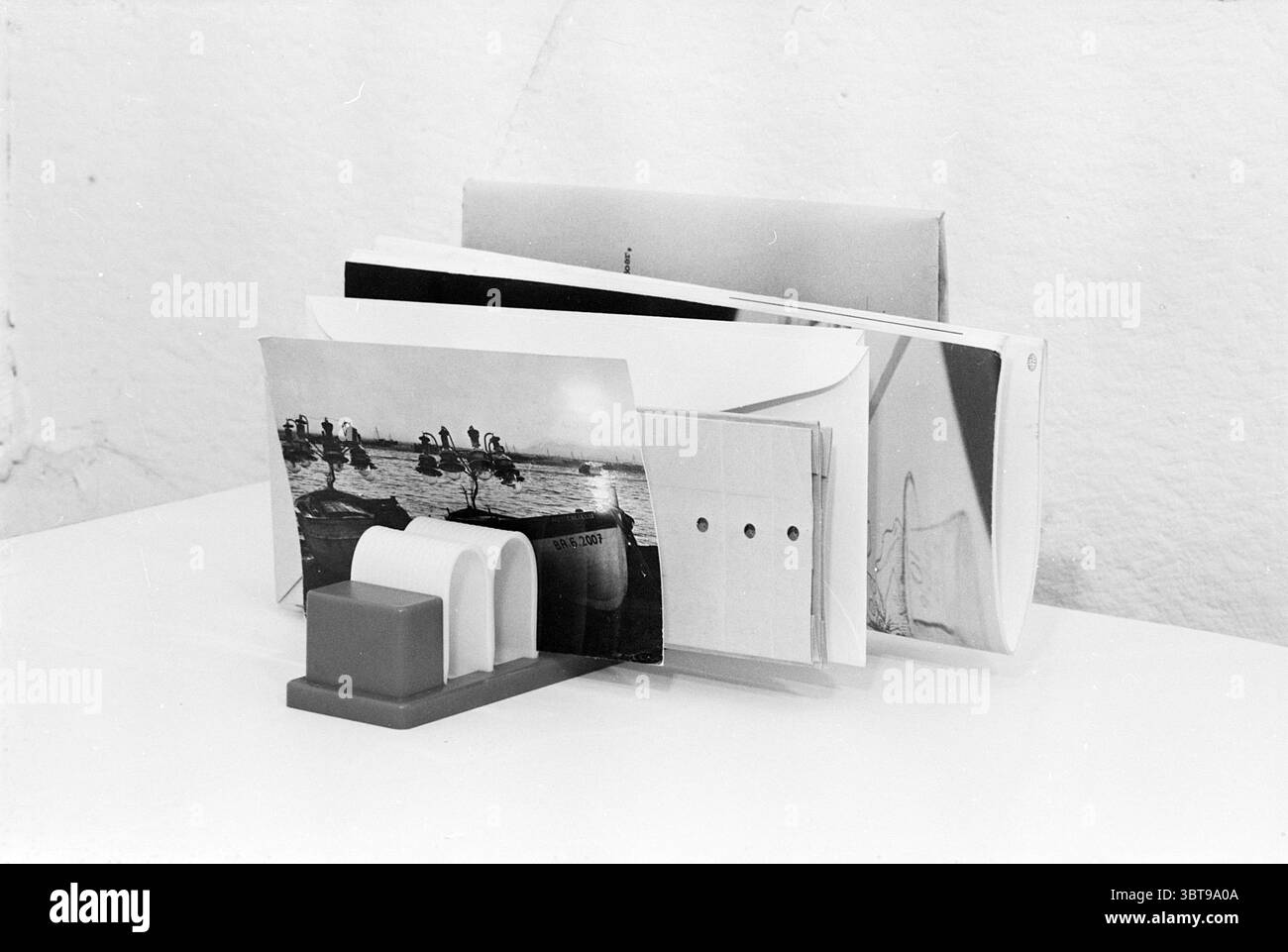 Invention Mr. Duynstee Inventions, Whizgle News, Dutch Desk, Niederlande, 1950 - 2000 am 26-07-1967. Dies sind die Themen im Bild. Die Szene zeigt eine Sammlung verschiedener Papiergegenstände, die ordentlich auf einem sauberen, weißen Tisch angeordnet sind. An der Vorderseite befindet sich ein rechteckiger Halter, der sich durch ein minimalistisches Design und eine weiche, gedämpfte Farbpalette auszeichnet. Der Halter unterstützt zwei Papierstapel neben einer separaten, größeren Karte. Die größte Karte zeigt ein Schwarzweiß-Foto mit Figuren, die möglicherweise an einer Aktivität in einem Gewässer beteiligt sind. Die Töne sind scharf, aber satt und betonen t Stockfoto