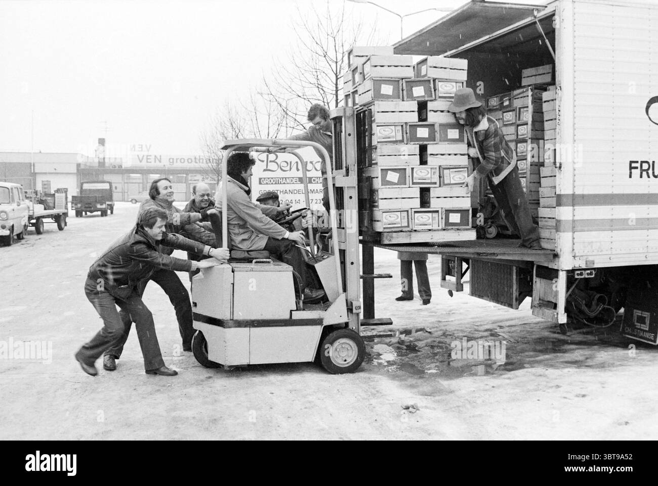 Rutschige Bedingungen: Entladen von Wagen bei der Gemüseauktion Winter, Whizgle News, Dutch Desk, Niederlande, 1950 - 2000 am 30-01-1979. Dies sind die Elemente im Bild. Die Szene fängt eine geschäftige Umgebung im Freien ein, wahrscheinlich im Winter in einem Gewerbegebiet. In der Mitte befindet sich ein weißer Gabelstapler, der neben einem großen Lkw geparkt ist, der teilweise mit ordentlich gestapelten Kisten beladen ist. Die Kartons sind beschriftet und scheinen systematisch angeordnet zu sein, was darauf hindeutet, dass sie wertvolle Güter enthalten. Mehrere Personen sind aktiv am Entladeprozess beteiligt. Einige Personen sind um den Gabelstapler versammelt Stockfoto