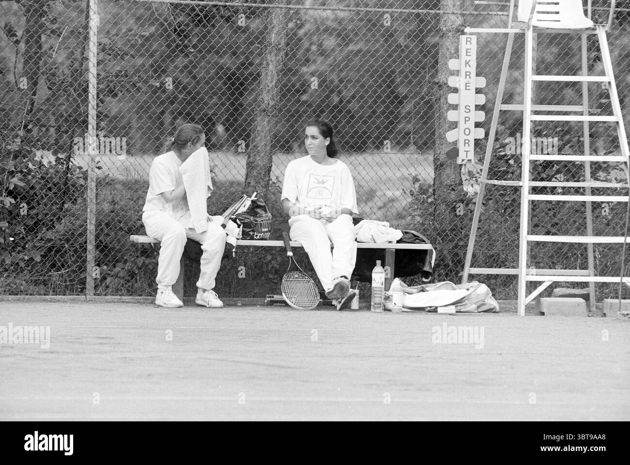 Tennisturnier in Bennebroek Bennebroek, Whizgle News, Dutch Desk, Niederlande, 1950 - 2000 am 22.06.1991. Diese Themen werden in der Abbildung angezeigt. Die Szene zeigt zwei Personen, die auf einer Bank neben einem Tennisplatz ruhen, was auf eine Pause zwischen den Aktivitäten hindeutet. Beide sind in passender weißer Sportkleidung gekleidet, die ein Gefühl von Gleichmäßigkeit und Teamarbeit vermittelt. Eine Person ist teilweise verdeckt, vielleicht wischt sie ihr Gesicht mit einem Handtuch ab, was darauf hindeutet, dass sie sich selbst angestrengt hat. Die Bank ist einfach und funktional, aus Holz gefertigt und vor dem Hintergrund eines Kettenlais positioniert Stockfoto
