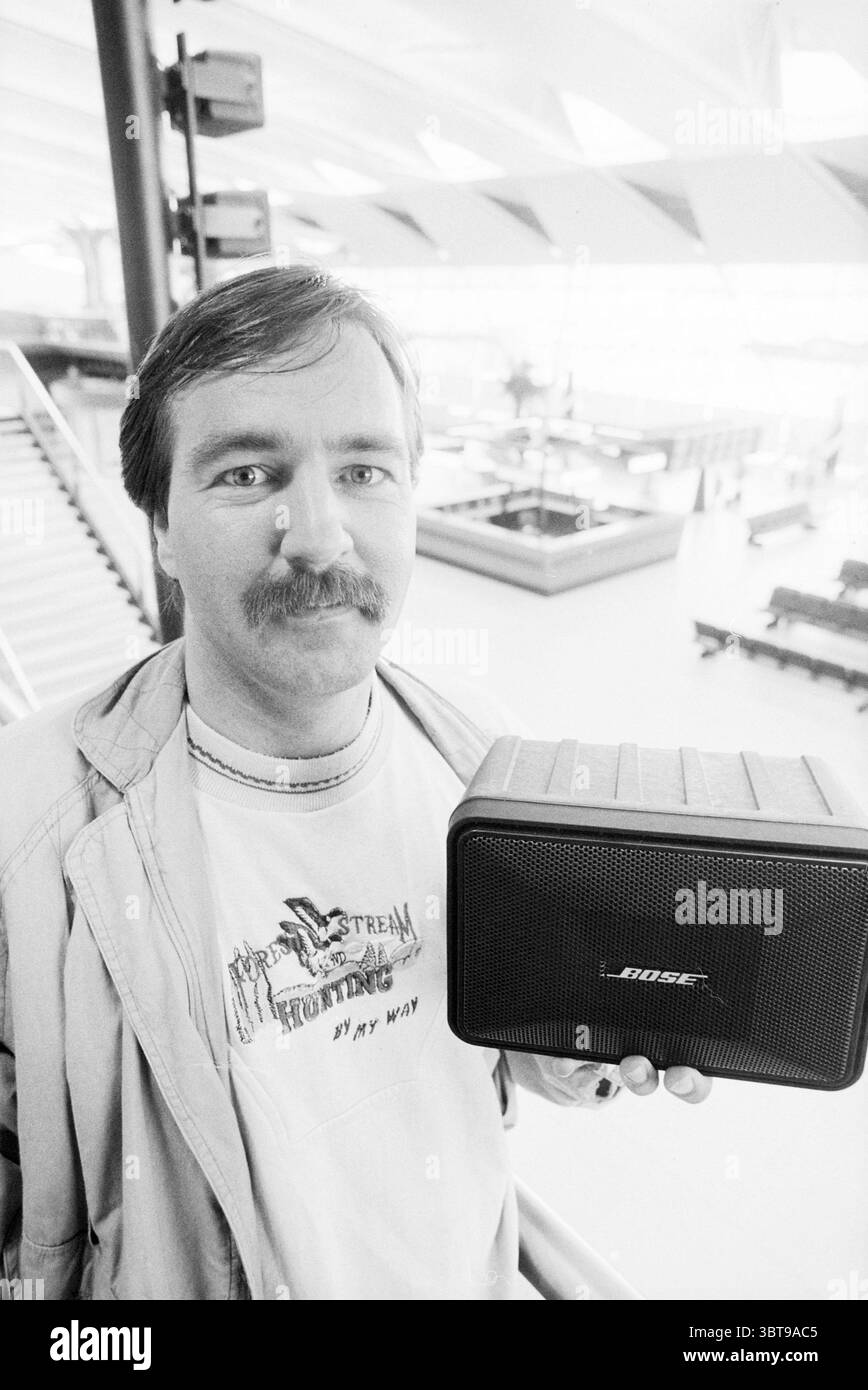 Arbeiter bei Schiphol Schiphol, Whizgle News, Dutch Desk, Niederlande, 1950 - 2000 am 02-05-1993. Das Bild enthält diese Themen. In einem geräumigen, modernen Innenbereich steht eine Person mit einem tragbaren Lautsprecher gut sichtbar. Die Szene vermittelt ein Gefühl von beiläufigem Selbstvertrauen. Die Person hat einen hellbraunen Schnurrbart und ein leicht zerzaustes Haar, gekleidet in einem bequemen T-Shirt, das mit einer Illustration versehen ist, die auf Themen wie Outdoor-Abenteuer oder persönlichen Ausdruck hinweist. Über dem T-Shirt sorgt eine leichte Jacke für einen mehrlagigen Look. Die Umgebung spiegelt einen modernen architektonischen Stil wider, der sich durch B Stockfoto