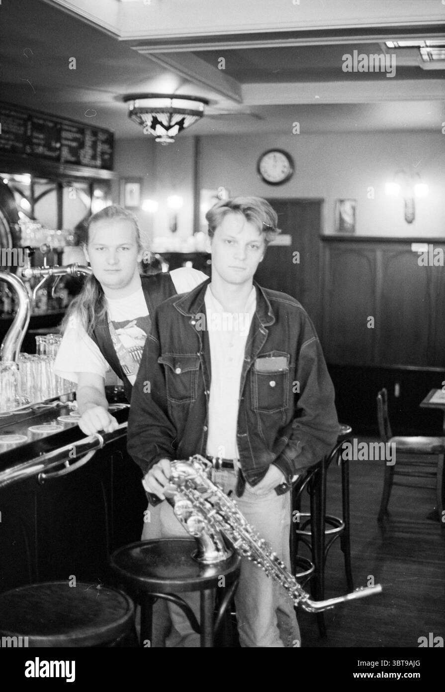 Merlijn Snitker & Xander Hubrecht, Whizgle News, Dutch Desk, Niederlande, 1950 - 2000 am 02-08-1993. Das Bild enthält diese Themen. In einer gemütlichen Einrichtung stehen zwei junge Männer in einer lebhaften Bar. Im Hintergrund befindet sich eine polierte Holzbar mit einer Reihe von Glaswaren, die ordentlich angeordnet sind. Ein Mann lehnt sich lässig an die Bar, eine selbstbewusste Haltung mit einem Arm leicht nach oben. Er hat lange Haare, die zurückgebunden sind, und trägt eine dunkle Weste über einem grafischen T-Shirt, das eine entspannte und dennoch stilvolle Atmosphäre andeutet. Der andere Mann steht nebenan, hält ein Saxophon nahe an seinem Körper und zeigt ein eher introspektives Stockfoto