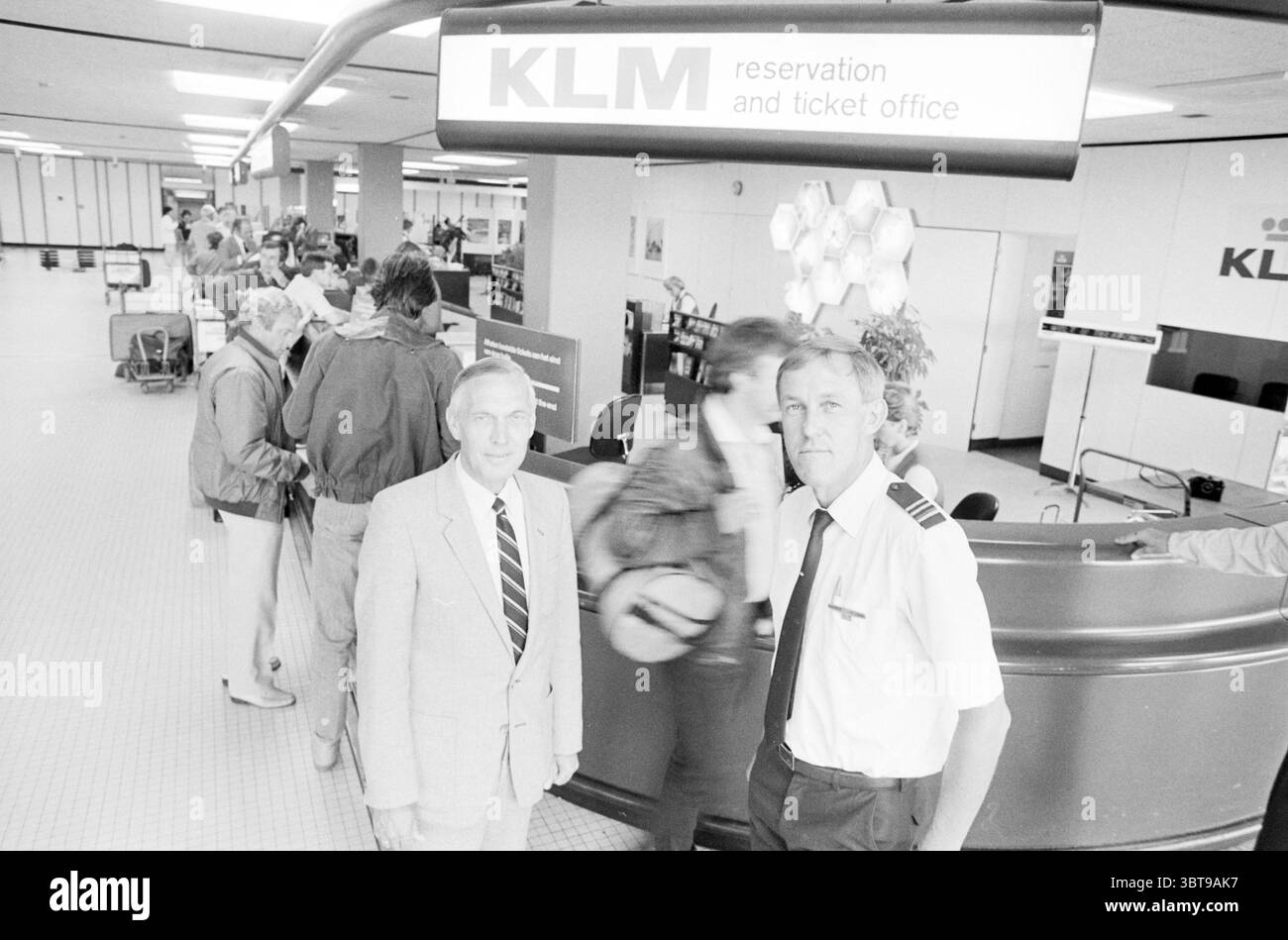 Ticketschalter KLM Schiphol Mr. Rensen und Scholte Schiphol Schiphol, Whizgle News, Dutch Desk, Niederlande, 1950 - 2000 am 27-06-1983. Diese Themen sind in der Abbildung dargestellt. Die Szene spielt in einem geräumigen Flughafenterminal mit viel Aktivität. Im Vordergrund stehen zwei Männer, einer in einem hellgrauen Anzug mit Krawatte und der andere in einer Pilotenuniform mit Schulterklappen. Sie stehen selbstbewusst in der Nähe des Check-in-Schalters, mit einem subtilen Lächeln, das auf ein Gefühl von Kameradschaft oder Professionalität hinweist. Hinter ihnen schlängelt sich eine Reihe von Reisenden in Richtung des Thekens, jede Person beschäftigt sich mit dem Stockfoto