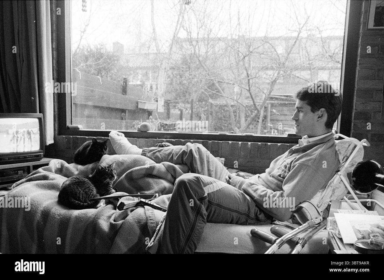 Der Eiskunstläufer Fausto de Marreiros erlitt bei einem Sturz eine Sehne und sieht jetzt Marathonläufer auf natürlichem Eis von seinem Bett aus., Whizgle News, Dutch Desk, Niederlande, 1950 - 2000 am 04-01-1992. Diese Themen sind in der Abbildung dargestellt. In einer gemütlichen Innenumgebung sitzt ein junger Mann bequem auf einer Couch, während sein Körper sich entspannt auf dem weichen Stoff entspannt. Er trägt ein helles Langarmhemd und eine lässige Hose, die eine entspannte Atmosphäre verkörpert. Seine Aufmerksamkeit richtet sich auf einen Fernsehbildschirm, der sanft leuchtet und ein sanftes Licht in den ansonsten gedämpften Raum wirft. Zwei Katzen Rollen sich neben ihm auf der Kuh Stockfoto