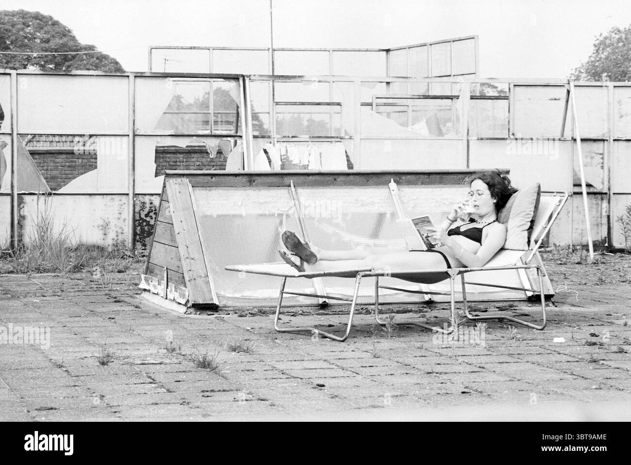 Bericht über temporäre Wohnräume in Haarlem (Frau im Bikini auf einer Liege auf einer großen Dachterrasse), Whizgle News, Dutch Desk, Niederlande, 1950 - 2000 am 21.06.1979. Diese Themen sind in der Abbildung dargestellt. Die Szene zeigt eine Frau, die auf einem Liegesessel liegt, in einem offenen, etwas verlassenen Bereich, der auf eine frühere Nutzung hindeutet, aber jetzt im Verfall ist. Sie trägt einen schwarzen Badeanzug, der einen Kontrast zu ihrer blassen Haut bildet, und ihr dunkles Haar ist locker gestylt und fällt um ihre Schultern. Die Frau wirkt entspannt, in ein Buch vertieft, was auf einen Moment der Freizeit hindeutet. Um sie herum, den Boden Stockfoto