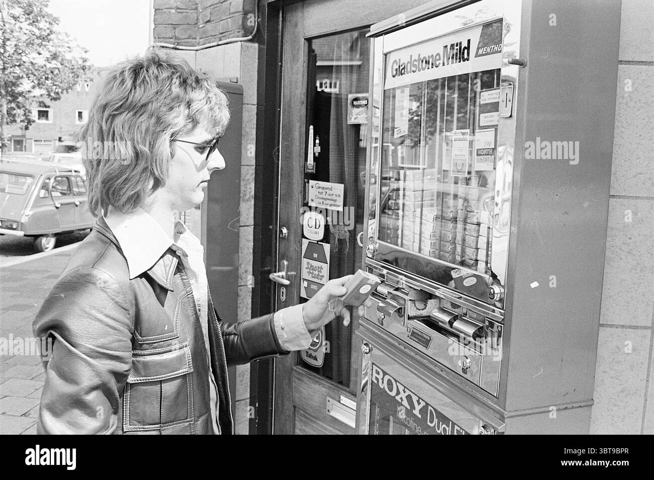 Zigarettenautomat, Whizgle News, Dutch Desk, Niederlande, 1950 - 2000 am 25-06-1974. Die Abbildung zeigt diese Themen. Ein junger Mann steht vor einem münzbetriebenen Automaten, der metallisch und leicht abgenutzt ist, was sein Alter widerspiegelt. Seine Frisur ist unverwechselbar und verfügt über lange, mehrschichtige Locken, die bis zu den Schultern reichen, was zu einem Retro-Vibe beiträgt. Er trägt eine dunkle Lederjacke mit silbernen Knöpfen, die über einem knusprigen Hemd mit Kragen und breitem Revers liegt und auf Modetrends aus der Vergangenheit hinweist. Die Kulisse zeigt eine Mischung aus urbaner Architektur, mit Ziegelmauern und großen Flächen Stockfoto