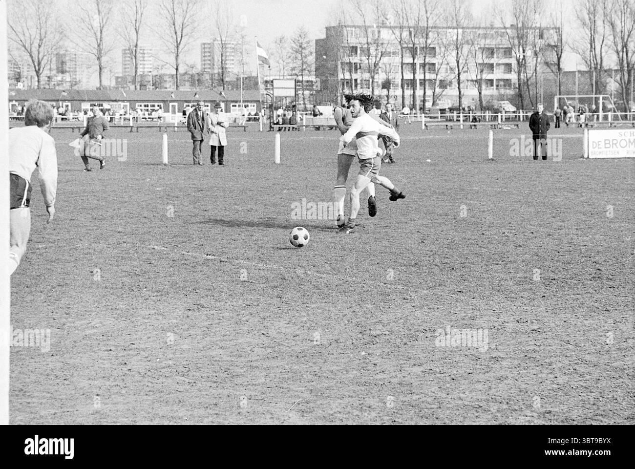 Kennemeren - A.D.O. Football, Whizgle News, Dutch Desk, Niederlande, 1950 - 2000 am 19.04.1970. Das Bild enthält diese Themen. Die Szene zeigt ein Fußballspiel im Freien auf einem grasbewachsenen Feld. Die Landschaft wird von einem reichen, erdigen Braun des Rasens dominiert, das leicht abgenutzt und von früheren Spielen markiert ist. In der Ferne ragt eine Reihe von hohen Gebäuden in den Himmel und bildet eine schroffe Kulisse vor dem ansonsten offenen Raum. Die Spieler, die in verschiedenen Trikots gekleidet sind, sind auf dem Spielfeld verstreut. Ein Spieler, der ein helles Trikot trägt, ist mitten im Geschehen und macht möglicherweise einen Pass oder versucht einen Schuss. Stockfoto