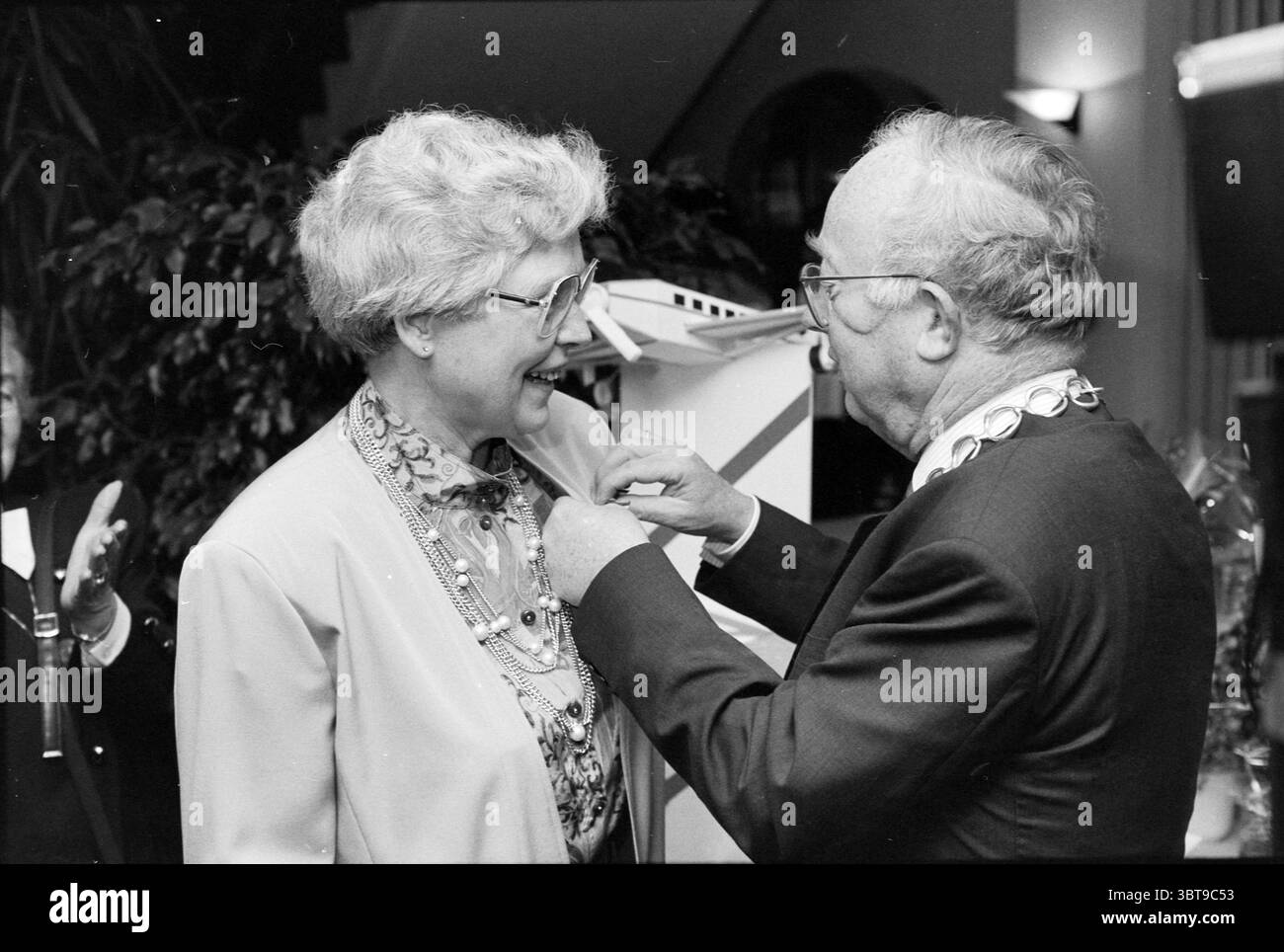 Königliche Dekoration Frau aus Sluisdam, Whizgle News, Dutch Desk, Niederlande, 1950 - 2000 am 16-01-1992. Die Abbildung zeigt diese Themen. In einem warm beleuchteten Innenbereich verbringen zwei Personen einen Moment der Feier. Die Atmosphäre fühlt sich fröhlich und intim an. Auf der linken Seite trägt eine Frau mit lockigem, silbernem Haar eine Brille und einen hellen Blazer über einer gemusterten Bluse, die Wärme und Zugänglichkeit ausstrahlt. Sie lächelt weit, ihr Ausdruck strahlt Glück und Aufregung aus. Rechts, ein älterer Mann in einem dunklen Anzug, der mit einer zeremoniellen Kette verziert ist, legt einen Bruder sanft fest Stockfoto