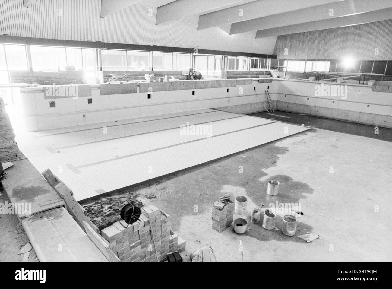 Bauschwimmbad., Whizgle News, Dutch Desk, Niederlande, 1950 - 2000 im Jahr 1978. Diese Themen werden in der Abbildung angezeigt. Die Szene zeigt einen weitläufigen Innenraum, der wie ein Schwimmbadbereich aussieht und derzeit im Bau oder Renovierung ist. Der Pool selbst ist groß, mit Betonwänden, die um ihn herum ragen, was auf ein modernes architektonisches Design hinweist. Die Oberfläche des Pools ist mit eleganten, weißen Fliesen bedeckt, die eine saubere und polierte Oberfläche vermuten lassen und darauf warten, dass Wasser seine Tiefe füllt. Um den Pool herum sind verschiedene Baumaterialien auf dem Boden verstreut, einschließlich Stockfoto
