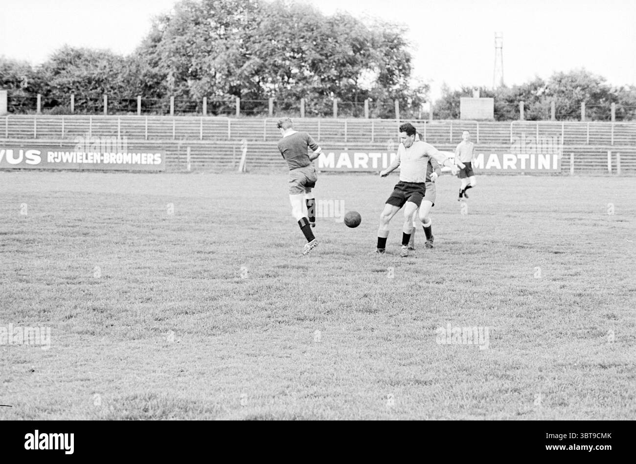 Justice versus Lawyers Football, Whizgle News, Dutch Desk, Niederlande, 1950 - 2000 am 09-06-1964. Das Bild enthält diese Themen. Auf einem grasbewachsenen offenen Feld spielen zwei Personen ein aktives Fußballspiel. Die Landschaft zeichnet sich durch üppiges, leuchtendes grünes Gras aus, das im Kontrast zu den monochromatischen Tönen der Komposition steht. Ein Spieler, der in einem hellen Trikot gekleidet ist, ist bereit, den Ball zu kicken, während ein anderer, der ein dunkleres Oberteil trägt, bereit zu sein scheint, abzufangen. Ihre Beine sind dynamisch positioniert, was die Bewegung und die Intensität des Spiels betont. Die Kugel befindet sich auf t Stockfoto