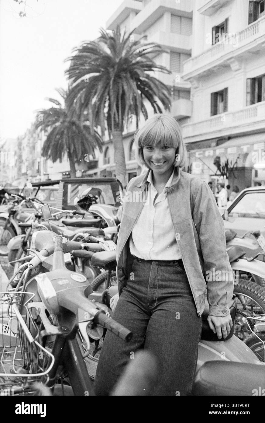 Reisen Sie Ibiza von Haarlems Dagblad in Zusammenarbeit mit Entspannungsreisen mit IMCA Marina Performance, Whizgle News, Dutch Desk, Niederlande, 1950 - 2000 am 02-04-1980. Dies sind die Themen im Bild. Im Vordergrund sitzt eine junge Frau lässig auf einem Vintage-Roller, ihr Verhalten ist warm und einladend, während sie ein helles Lächeln strahlt. Sie hat schulterlange, glatte Haare mit einem hellen blonden Farbton, eingerahmt von einer schlichten grauen Jacke, die ihr einen entspannten und doch stylischen Look verleiht. Ihr Outfit wird durch eine hochgeschnittene Jeans aus dunklem Denim abgerundet, die die zeitlose Ästhetik der Szene unterstreicht. Um ihn herum Stockfoto