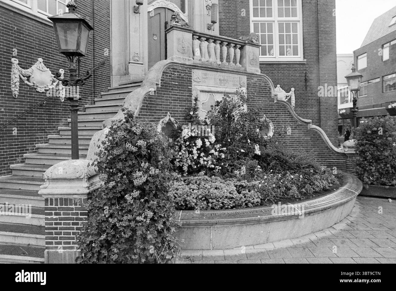Blumenbeet vor dem Rathaus Z'voort Zandvoort, Whizgle News, Dutch Desk, Niederlande, 1950 - 2000 am 07-06-1995. Das Bild enthält diese Themen. Die Szene fängt eine architektonische Umgebung mit komplizierten Details und einem Sinn für Geschichte ein. Eine große Treppe, flankiert von kunstvollen Geländern, führt zu einem wunderschön gestalteten Eingang. Die Stufen bestehen aus Stein, deren Oberfläche Anzeichen von Verschleiß aufweist, was auf einen langen Lauf der Zeit hindeutet. Vor dem Treppenhaus fällt üppiges Grün über ein rundes Blumenbeet, das von einer Vielzahl von Pflanzen in voller Blüte überschwemmt wird. Farbtöne von wh Stockfoto