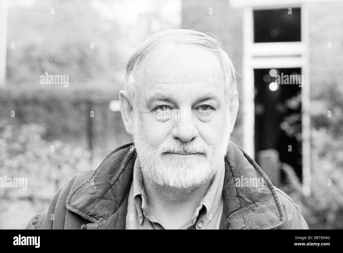 Eric Carle Kinderbuchautor Bücher und Buchhändler People Writing, Whizgle News, Dutch Desk, Niederlande, 1950 - 2000 am 11-10-1988. Dies sind die Elemente im Bild. Die Szene zeigt einen älteren Mann mit einem vollen, grauen Bart und leicht zerzausten Haaren, der ein Gefühl von Weisheit und Erfahrung vermittelt. Sein Ausdruck ist ruhig und besinnlich, mit einem Hauch von Wärme in seinen Augen. Er trägt eine dunkelfarbene Jacke mit einer robusten Struktur, die einen praktischen, bodenständigen Stil andeutet. Der Hintergrund zeigt Hinweise auf ein Gebäude mit warmen Wänden, die teilweise von Laub verdeckt sind und eine organische Struktur schaffen Stockfoto