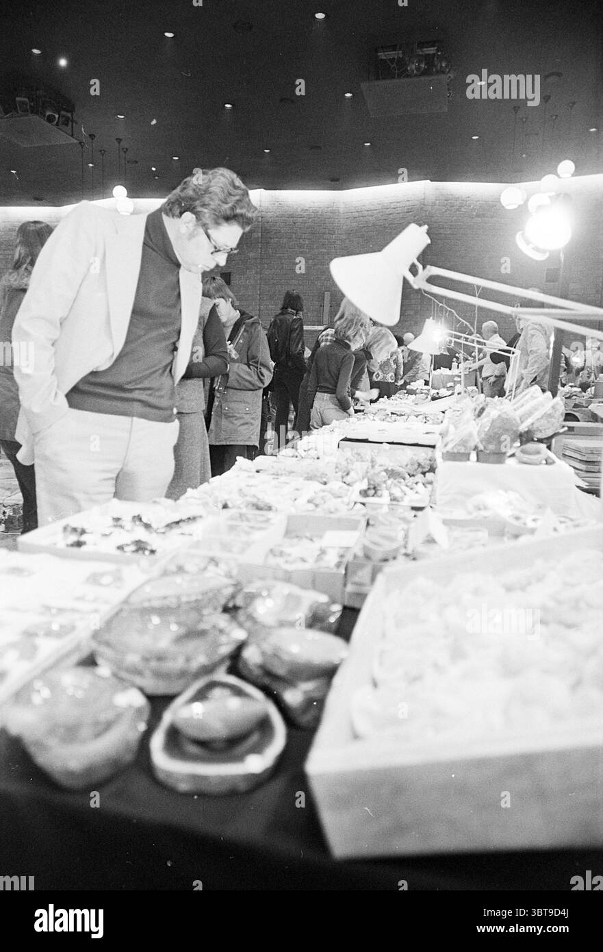 Tag der Geologen im Schouw Velsen Noord Geology Velsen-Noord Bleyenhoevelaan, Whizgle News, Dutch Desk, Niederlande, 1950 - 2000 am 20-03-1976. Dies sind die Elemente im Bild. Die Szene fängt einen belebten Indoor-Markt mit einer Vielzahl von farbenfrohen Gegenständen auf den Tischen ein. Im Vordergrund sind verschiedene handwerkliche Lebensmittel sorgfältig angeordnet, mit lebendigen Früchten, ordentlich verpackten Snacks und verlockenden Backwaren. Die Texturen der Artikel reichen von glänzenden Oberflächen mit glänzenden Früchten bis hin zum rustikalen Aussehen von Brot und Gebäck, was ein Augenschmaus schafft. Ein gut gekleidetes M Stockfoto