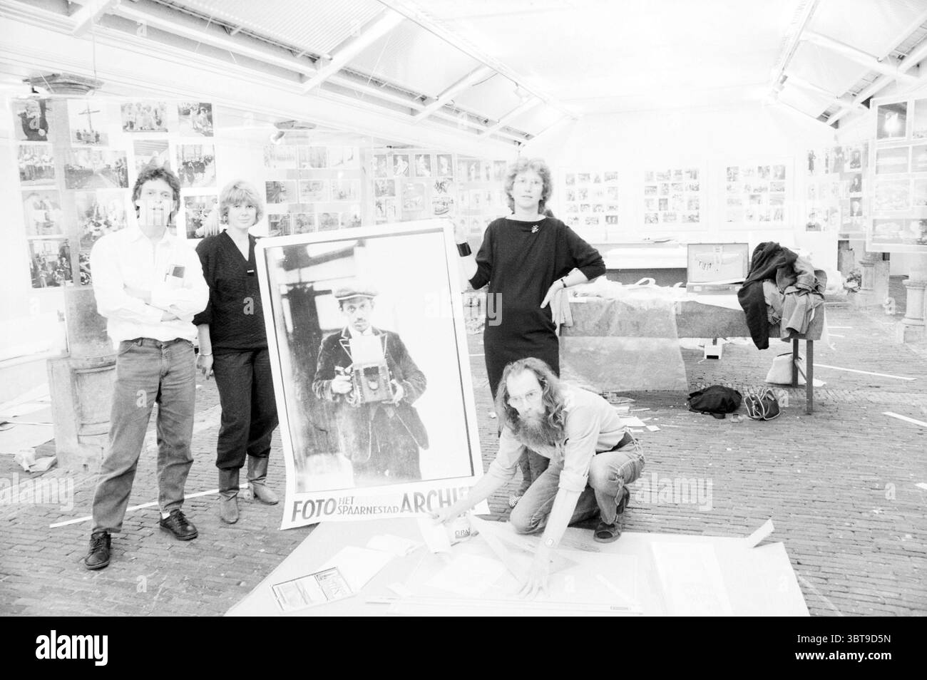Organisation einschließlich Harm Botman Fotoausstellung Spaarne Archive Photography, Whizgle News, Dutch Desk, Niederlande, 1950 - 2000 am 26-08-1986. Dies sind die Themen im Bild. In einer geräumigen, gut beleuchteten Galerie versammeln sich vier Personen um ein großes, Vintage-Foto, das eine Person mit einer Kamera zeigt. Die Atmosphäre ist kollaborativ und kreativ, denn künstlerische Projekte und Fotografie sind überall im Raum verstreut. Die erste Person links, die ein weißes Langarmhemd und eine dunkle Hose trägt, hat einen engagierten Ausdruck, der möglicherweise eine Idee oder ein Konzept erklärt. Ne Stockfoto