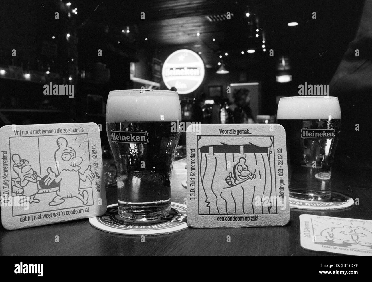 Ziemlich sichere Biermatten, Whizgle News, Dutch Desk, Niederlande, 1950 - 2000 am 02-04-1992. Die Abbildung zeigt diese Themen. Die Szene spielt in einer gemütlichen Bar mit warmer, dezenter Beleuchtung, die ein sanftes Leuchten über die Holzmöbel wirft. Im Vordergrund sitzen zwei Gläser Schaumbier auf einer polierten Barfläche, deren goldene Flüssigkeit unter dem Umgebungslicht glitzert. Der Schaum auf der Oberseite ist dick und cremig und setzt sich langsam ein, als würde er zu einem Schluck einladen. Die Brille wird von zwei Untersetzern mit skurrilen Illustrationen eingerahmt. Die eine stellt einen skurrilen Charakter mit übertriebenen Merkmalen dar, die andere Stockfoto