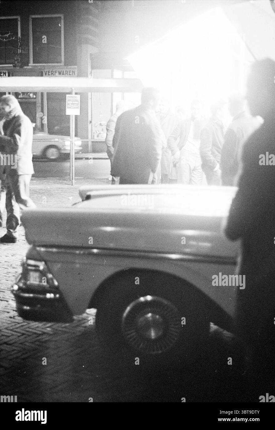 Straßenszene. Autohandel Haarlem Botermarkt Niederlande, Whizgle News, Dutch Desk, Niederlande, 1950 - 2000 am 07-11-1963. Die Abbildung zeigt diese Themen. In einer belebten urbanen Szene steht ein Oldtimer im Mittelpunkt, der leicht links geparkt ist. Sein Körper ist in einer sanften Creme lackiert und mit Chromakzenten versehen, die das Licht auffangen und die abgerundeten Konturen und das klassische Design hervorheben. Das Fahrzeug nimmt eine herausragende Position ein und schafft einen faszinierenden Blickwinkel vor dem Hintergrund. Um das Fahrzeug herum steht eine Gruppe von Personen, die sich mit Gesprächen beschäftigen. Sie sind in dunklen, gedämpften Tönen gekleidet, Stockfoto