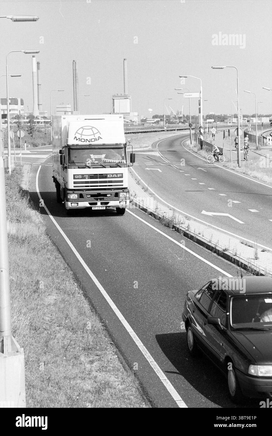 Kreuzung zwischen de Geul und Kanaalstraat. IJmuiden the Netherlands, Whizgle News, Dutch Desk, Niederlande, 1950 - 2000 am 08-1991. Das Bild enthält diese Themen. Die Szene zeigt eine befahrene Straße, die durch eine sanft gewundene Kurve gekennzeichnet ist, die in die Distanz führt. Im Vordergrund dominiert ein großer Lkw auf der linken Seite, der in einem hellen weiß mit fetten Buchstaben auf der Seite gestrichen ist, die wahrscheinlich auf einen Firmennamen hinweisen. Seine Größe erzeugt ein Gefühl von Präsenz, da er die Spur beherrscht, die er einnimmt. Rechts fährt ein kleineres Auto neben dem Lkw, das sich farblich mit dunkleren Schattierungen abhebt Stockfoto