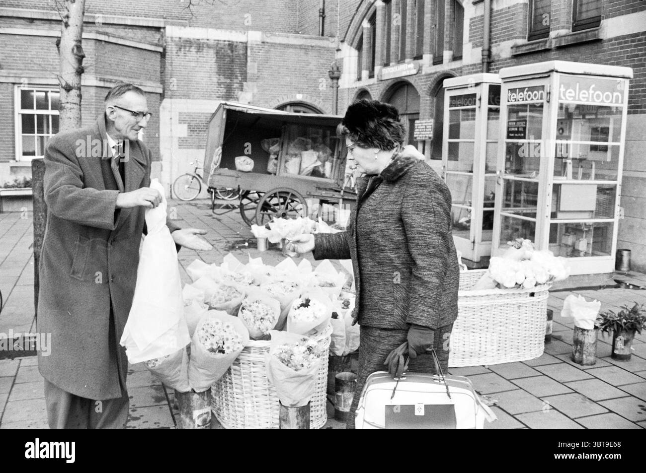 Stationsplein Haarlem Stationsplein The Netherlands, Whizgle News, Dutch Desk, Niederlande, 1950 - 2000 am 01-11-1962. Das Bild enthält diese Themen. Die Szene entfaltet sich in einem Open-Air-Markt, der durch seine zwanglose, einladende Atmosphäre gekennzeichnet ist. Zwei Personen führen einen lebhaften Austausch. Ein Mann, gekleidet mit dunklem Mantel und Brille, steht links. Er scheint animiert zu sein, während er einen umwickelten Blumenstrauß hält und wahrscheinlich Gespräche über die Auswahl führt. Nach rechts neigt sich eine Frau in einem stilvollen, dunklen Mantel und trägt eine schicke Frisur leicht nach vorne. Stockfoto