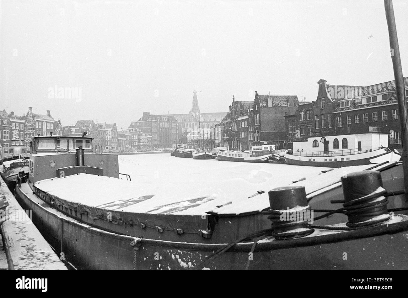 Winter in Haarlem Winter, Whizgle News, Dutch Desk, Niederlande, 1950 - 2000 auf 31-01-1972. Diese Themen werden in der Abbildung angezeigt. Die Szene zeigt eine ruhige, winterliche Kanalkulisse, in der das Wasser teilweise gefroren ist und eine glatte, weiße Oberfläche schafft, die die umliegenden Gebäude reflektiert. Im Vordergrund befindet sich ein robustes Boot mit einem abgerundeten Rumpf, der mit einer Schneeschicht bedeckt ist. Die satten Farben stehen im Kontrast zum hellen weiß und verleihen dem Bild mehr Tiefe. Im Hintergrund säumen mehrere historische Gebäude den Kanal, die durch komplizierte Giebeln und enge Fenster gekennzeichnet sind. Ihre Fassaden sind stumm Stockfoto