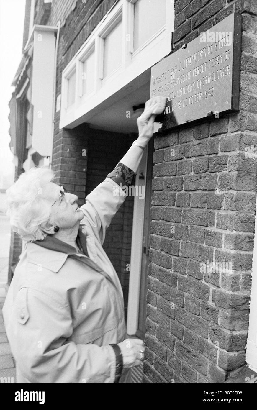 Frau Hart in Verbindung mit Plaque Haarlem the Netherlands, Whizgle News, Dutch Desk, Niederlande, 1950 - 2000 am 03-02-1993. Diese Themen werden in der Abbildung angezeigt. In einer gedämpften urbanen Umgebung greift eine Frau mit einer strukturierten Wand in Berührung, wobei ihre Aufmerksamkeit auf ein Objekt gerichtet ist, das daran befestigt ist. Sie ist in einen hellen Mantel gekleidet, der sich sanft mit dem dunkleren Backstein des Gebäudes abhebt. Ihr Haar ist weiß und ordentlich gestylt, und sie scheint ein fortgeschrittenes Alter zu sein und zeigt ein Gefühl von Weisheit und Erfahrung. Die Wand selbst hat satte, dunkelbraune Töne, mit einzelnen Ziegeln sichtbar, die Tiefe verleihen Stockfoto