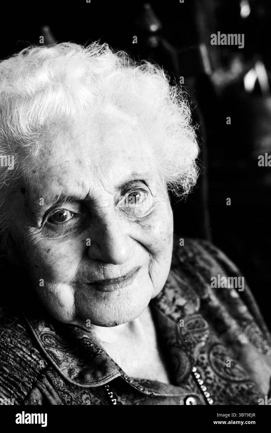 Mrs. Woltman 100 Jahre alt. Moerberg IJmuiden IJmuiden the Netherlands, Whizgle News, Dutch Desk, Niederlande, 1950 - 2000 auf 31-03-1997. Das Bild enthält diese Themen. In einem schwach beleuchteten Raum nimmt eine ältere Frau den Vordergrund ein, ihr ausdrucksstarkes Gesicht leuchtet sanft und erzeugt eine Wärme, die sich im Kontrast zu den umgebenden Schatten befindet. Ihr Haar, eine Wolke aus weißen Locken, umrahmt ihr Gesicht sanft, während ihre Haut die zarten Zeichen des Alters zeigt – subtile Linien, die Geschichten eines langen Lebens erzählen. Sie blickt direkt auf den Betrachter, ihre großen Augen funkeln vor Weisheit und Neugier. Das Zimmer Stockfoto