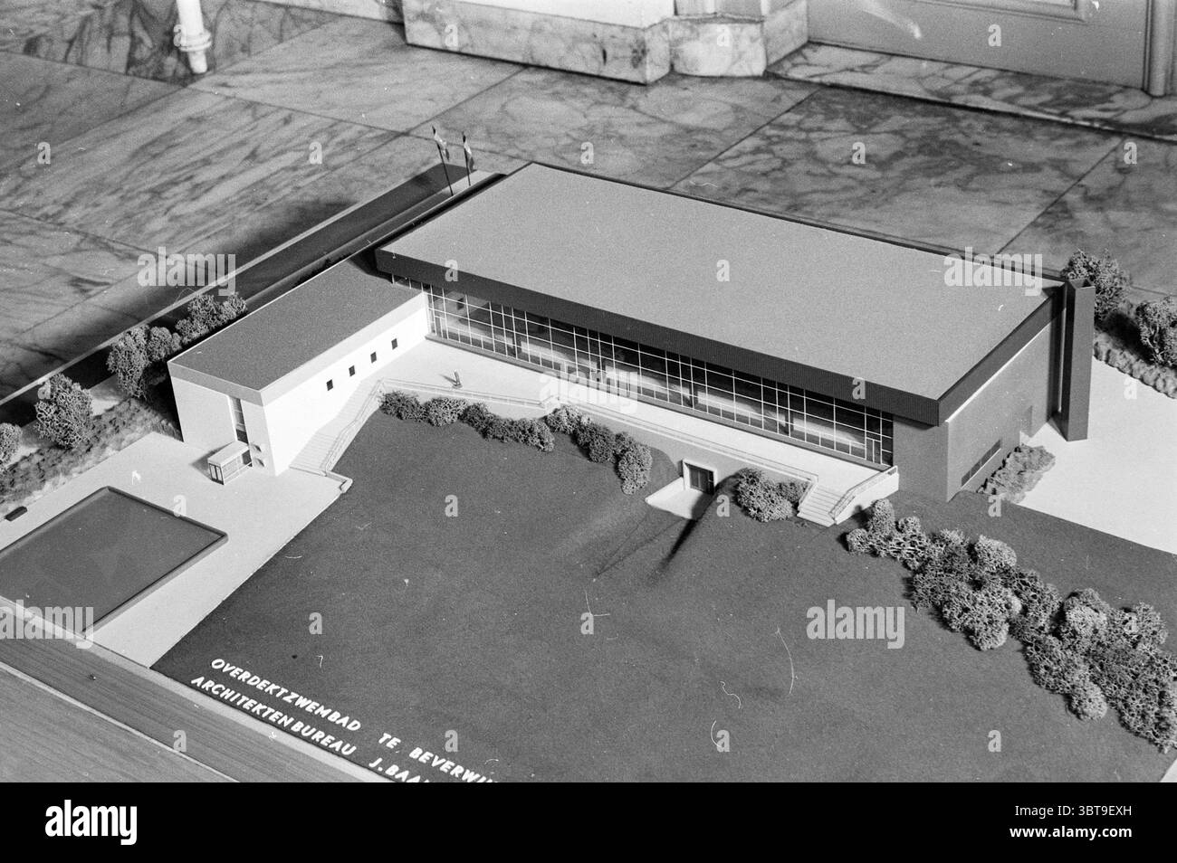 Beverwijk Swimming Pool Model, Whizgle News, Dutch Desk, Niederlande, 1950 - 2000 am 22-02-1963. Diese Themen werden in der Abbildung angezeigt. Die Szene zeigt ein detailliertes Architekturmodell, das deutlich sichtbar auf einer Ebenen Fläche angezeigt wird. Die Struktur selbst ist modern und minimalistisch, zeichnet sich durch klare Linien und geometrische Formen aus. Sie besticht durch ihre markante Glasfassade, die die angrenzende Umgebung reflektiert und ein Gefühl von Transparenz und Offenheit vermittelt. Um das zentrale Gebäude herum gibt es sorgfältig gepflegte Grünflächen, darunter Sträucher und kleine Bäume, die einen Touch verleihen Stockfoto