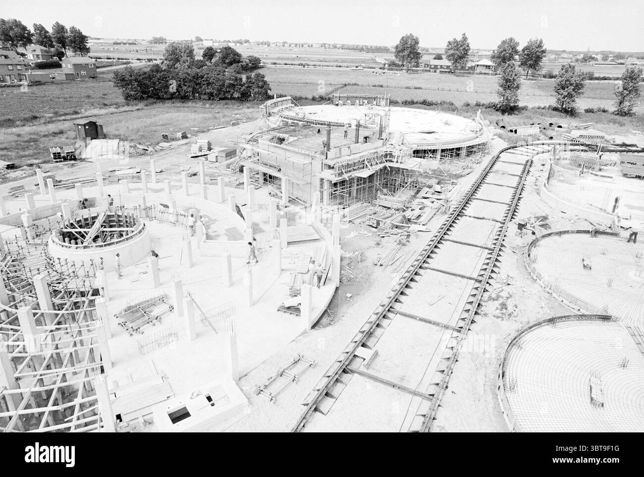 Bau der Wasseraufbereitungsanlage Zwaanshoek Baugruben Water Zwaanshoek, Whizgle News, Dutch Desk, Niederlande, 1950 - 2000 am 10-07-1979. Dies sind die Elemente im Bild. Die Szene zeigt eine weitläufige Baustelle, die durch eine Vielzahl von laufenden Bautätigkeiten gekennzeichnet ist. Im Zentrum der Komposition stehen zwei große, teilweise geformte Strukturen mit Skelettgerüsten aus Stahl und Beton, die sich gegen den Horizont erheben. Diese Bauwerke sind von Gerüsten umgeben, was darauf hinweist, dass aktiv gearbeitet wird. Die Farbpalette ist überwiegend gedämpft, mit Schattierungen Stockfoto