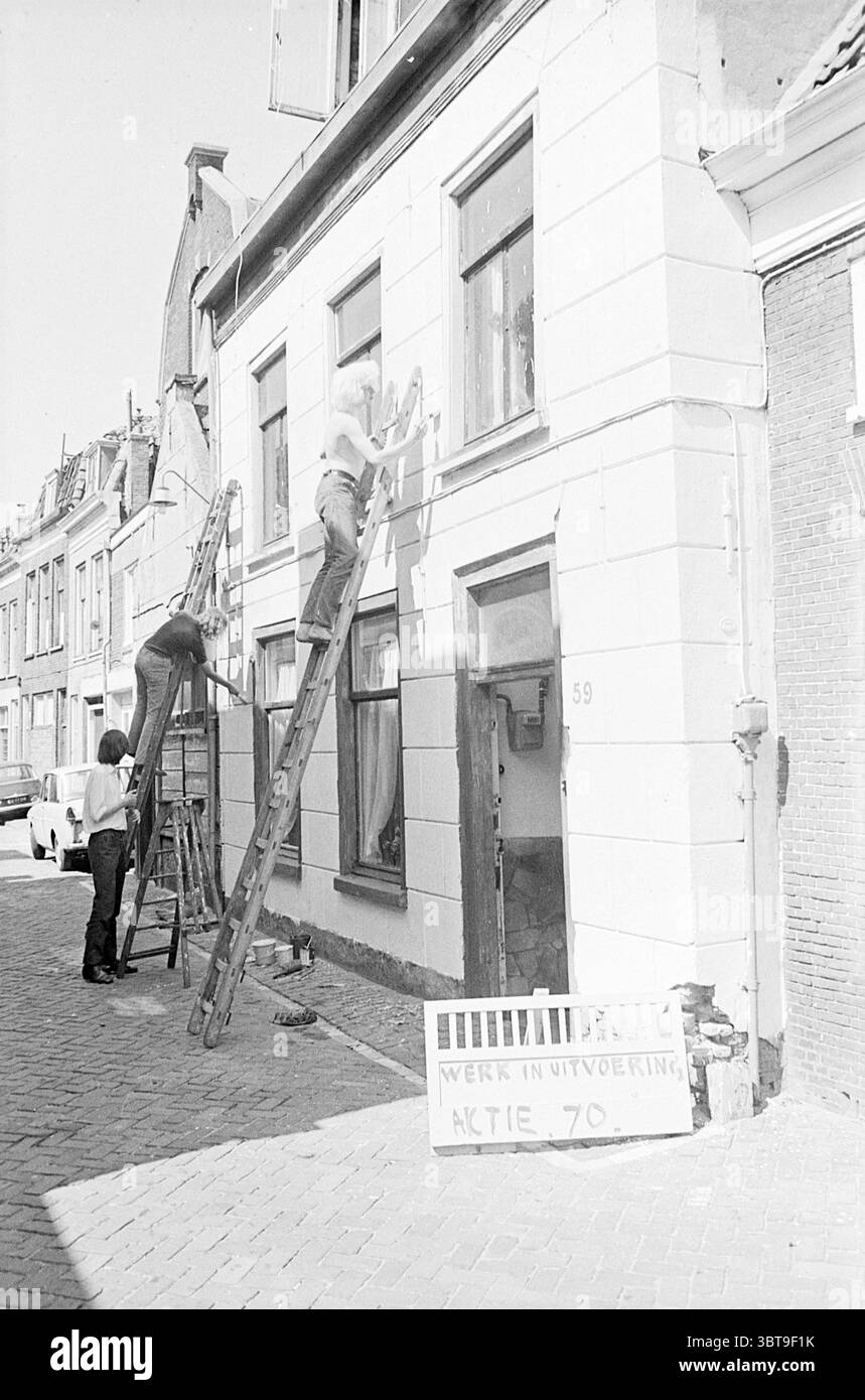 Gemälde eines Gebäudes aufgrund der Aktion 70., Whizgle News, Dutch Desk, Niederlande, 1950 - 2000. Das Bild enthält diese Themen. Die Szene fängt eine belebte Straße in einer malerischen, urbanen Umgebung ein. Der Schwerpunkt liegt auf zwei Mitarbeitern, die an der Sanierung der Fassade eines Gebäudes beteiligt sind. Eine Person sitzt auf einer hohen Leiter und trägt Farbe auf den oberen Teil der Wand auf, während eine andere auf einer unteren Leiter steht, um Materialien vorzubereiten oder bei der Arbeit zu helfen. Beide tragen Arbeitskleidung, was auf ein Gefühl von Fleiß hindeutet. Das Gebäude selbst weist einen klassischen architektonischen Stil auf. Stockfoto