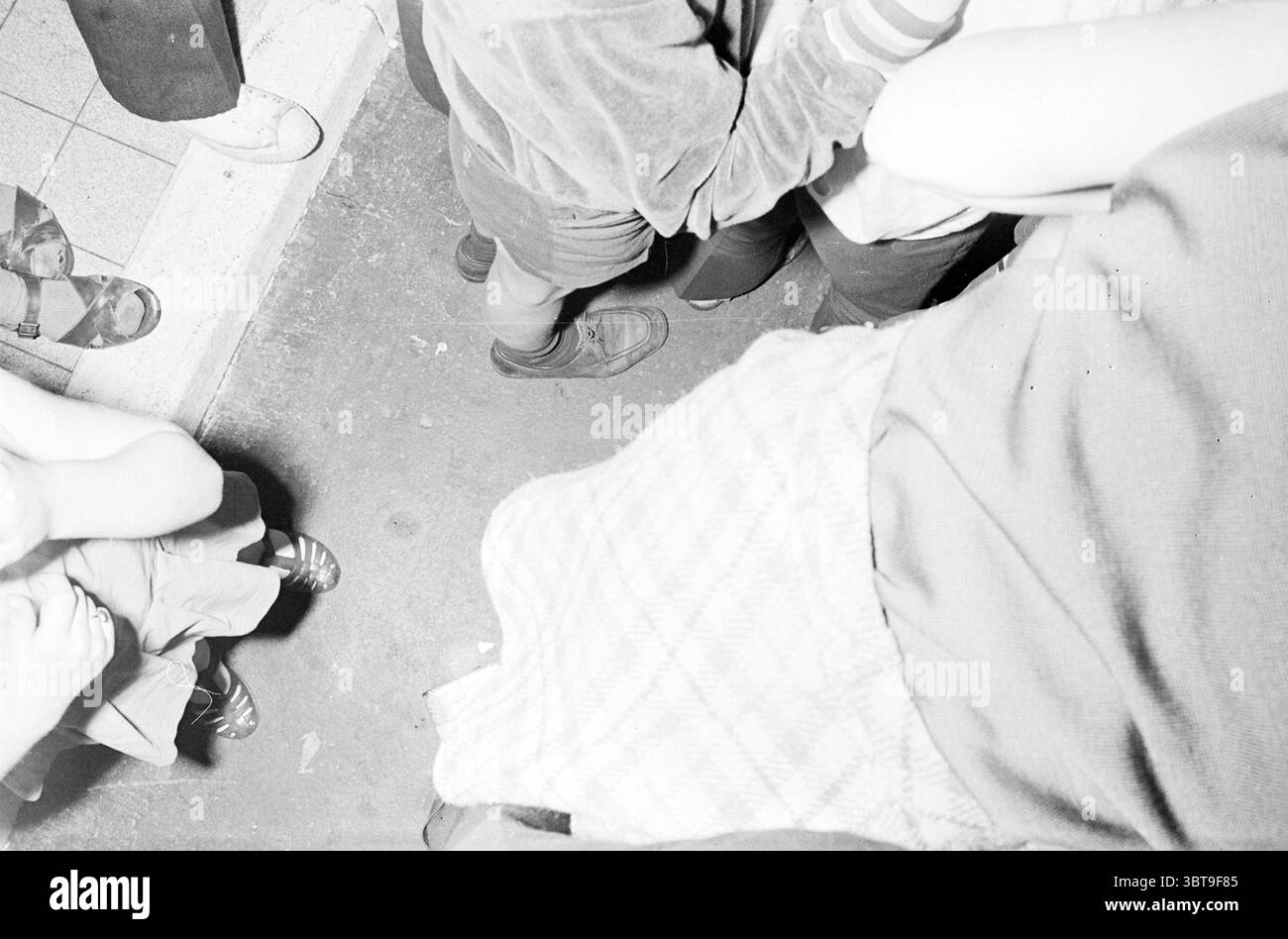 10000. Besucher Stuif Stuif Stuif Stuif, Whizgle News, Dutch Desk, Niederlande, 1950 - 2000 auf 30-07-1970. Diese Themen werden in der Abbildung angezeigt. Die Szene erfasst eine dynamische und eng geschnittene Ansicht einer Menge, bei der der Schwerpunkt hauptsächlich auf einer Kollektion von Gliedmaßen und Kleidung liegt. Die Farben sind überwiegend monochromatisch, wobei Grauschattierungen als Hintergrund fungieren. Dunklere Töne vermischen sich mit wärmeren Tönen, die auf unterschiedliche Stoffstrukturen hinweisen. Im Vordergrund befinden sich mehrere Paar Beine und Füße, von denen einige in formellen Schuhen gestopft sind, während andere eher lässige Sandalen oder Fußschuhe tragen Stockfoto