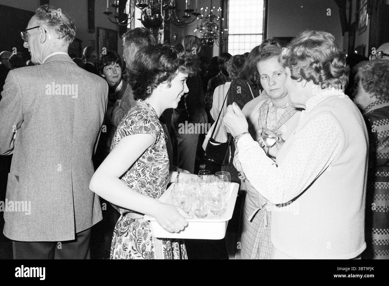 Abschiedsempfang Alderman Mensink Aldermen, Whizgle News, Dutch Desk, Niederlande, 1950 - 2000 am 29-04-1982. Diese Themen sind in der Abbildung dargestellt. Bei einem geschäftigen Treffen im Innenbereich steht eine junge Frau an der Spitze, die ein weißes Tablett mit kleinen Gläsern hält, in dem man wahrscheinlich Getränke servieren kann. Sie trägt ein gemustertes Kleid mit einem komplizierten Blumenmuster, das die gedämpfte Kleidung der Menge um sie herum kontrastiert. Ihr Haar ist in einer klassischen, für diese Zeit typischen Mode gestaltet, umrahmt ihr Gesicht und betont ein leichtes Lächeln. Um sie herum, engagieren sich verschiedene Menschen in Konv Stockfoto