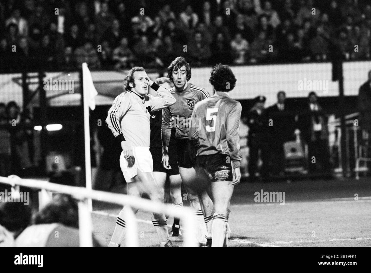 Belgien - Niederlande Fußball, Whizgle News, Dutch Desk, Niederlande, 1950 - 2000 am 19-11-1980. Diese Themen werden in der Abbildung angezeigt. Die Szene fängt einen dynamischen Moment auf einem Fußballfeld ein, gefüllt mit der Intensität eines Wettkampfes. Die Kulisse bietet ein Nachtspiel, beleuchtet von hellen Flutlichtern, die scharfe Kontraste zwischen den Spielern und den dunklen Ständen mit Zuschauern erzeugen, was eine aufgeladene Atmosphäre schafft. Im Vordergrund stehen drei Spieler. Einer, in einem hellen Jersey gekleidet, wirkt aufgeregt, mit einem animierten Ausdruck und einer mittigen Geste, während er sich einsetzt Stockfoto